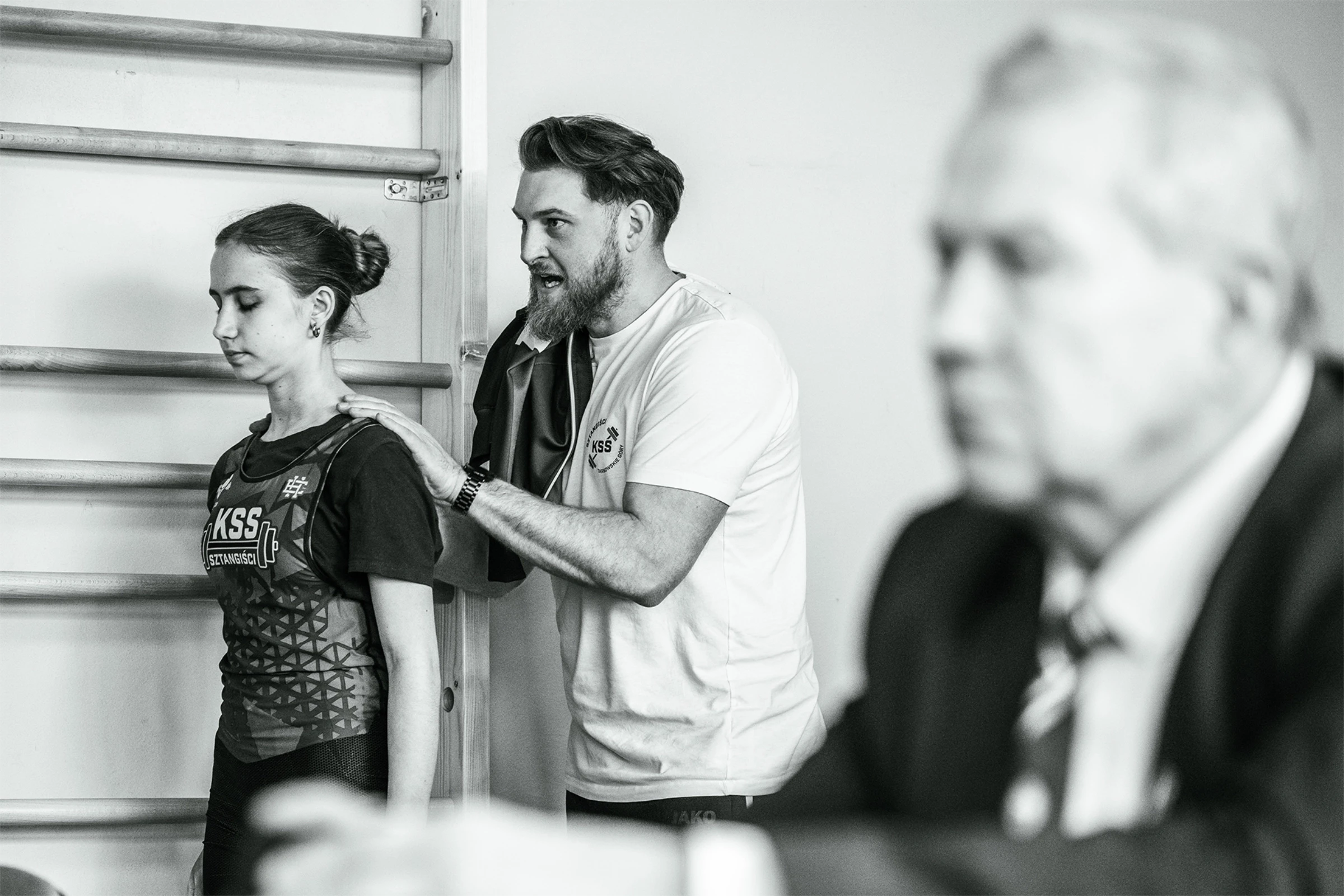 Czarno-białe zdjęcie trenera Sztangistów KSS Tarnowskie Góry udzielającego wsparcia zawodniczce przy drabinkach gimnastycznych.