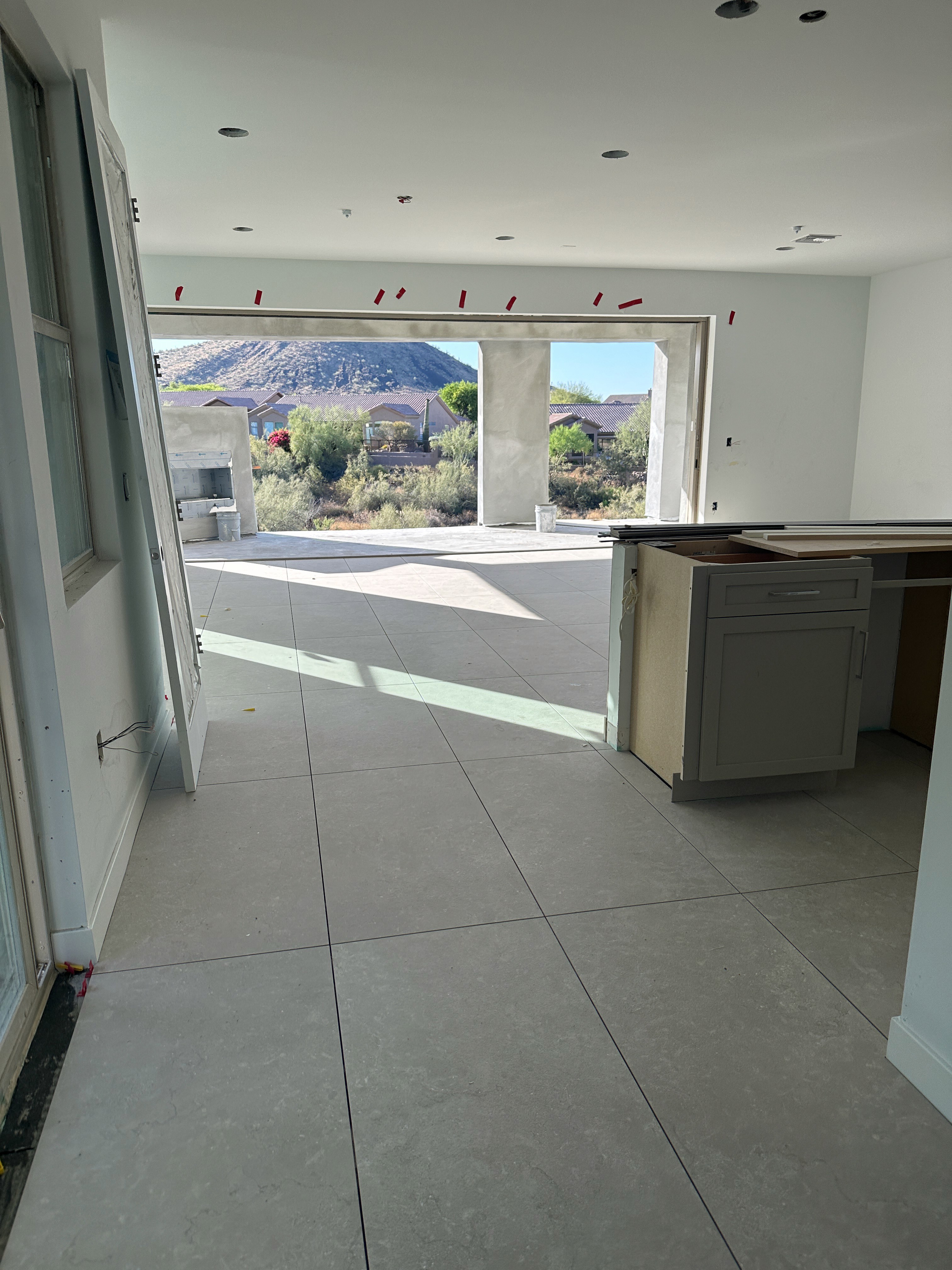 Modern open-plan living area under construction with large sliding glass doors, kitchen island, and unfinished tile flooring with a desert view.