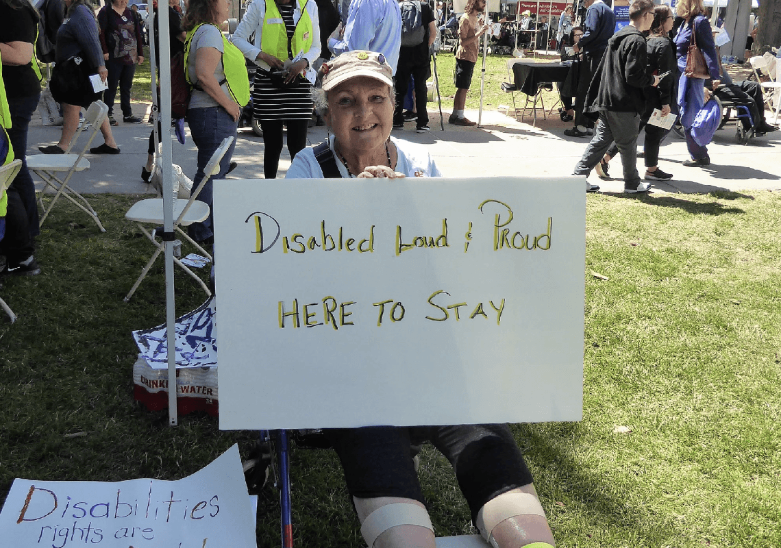 A woman seated outdoors holds a sign that reads, “Disabled Loud & Proud, HERE TO STAY,” smiling at the camera during a public event.