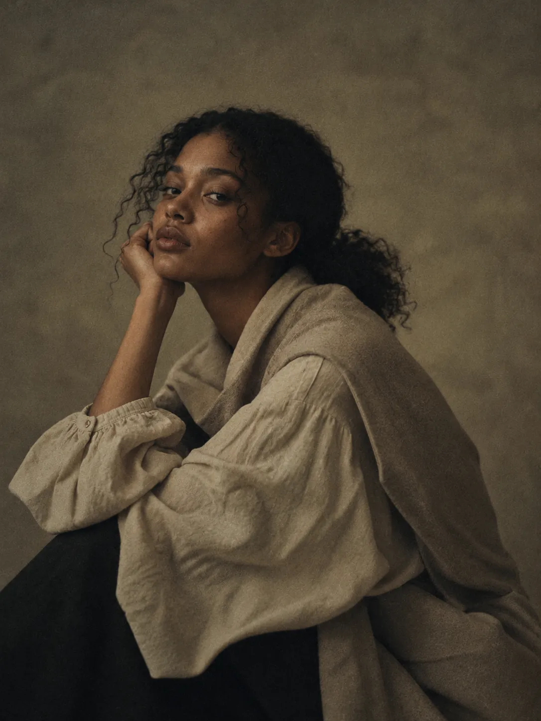 A young woman with curly hair resting her chin on her hand, wearing a loose linen shirt, against a plain mottled backdrop.