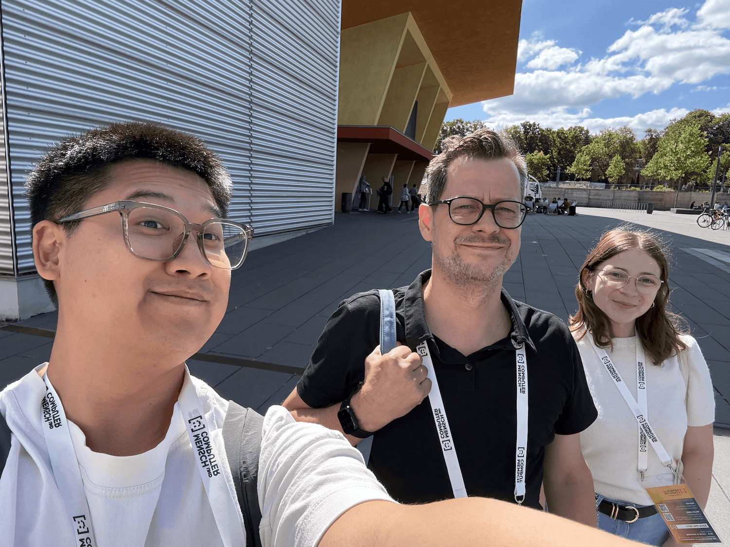 Drei Personen mit Konferenz-Ausweisen vor einem modernen Gebäude mit Metallfassade und orangefarbenem Dach bei sonnigem Himmel