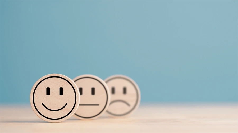 Three wooden smiley face icons representing feedback or satisfaction levels — happy, neutral, and sad — placed on a light surface with a blue background.
