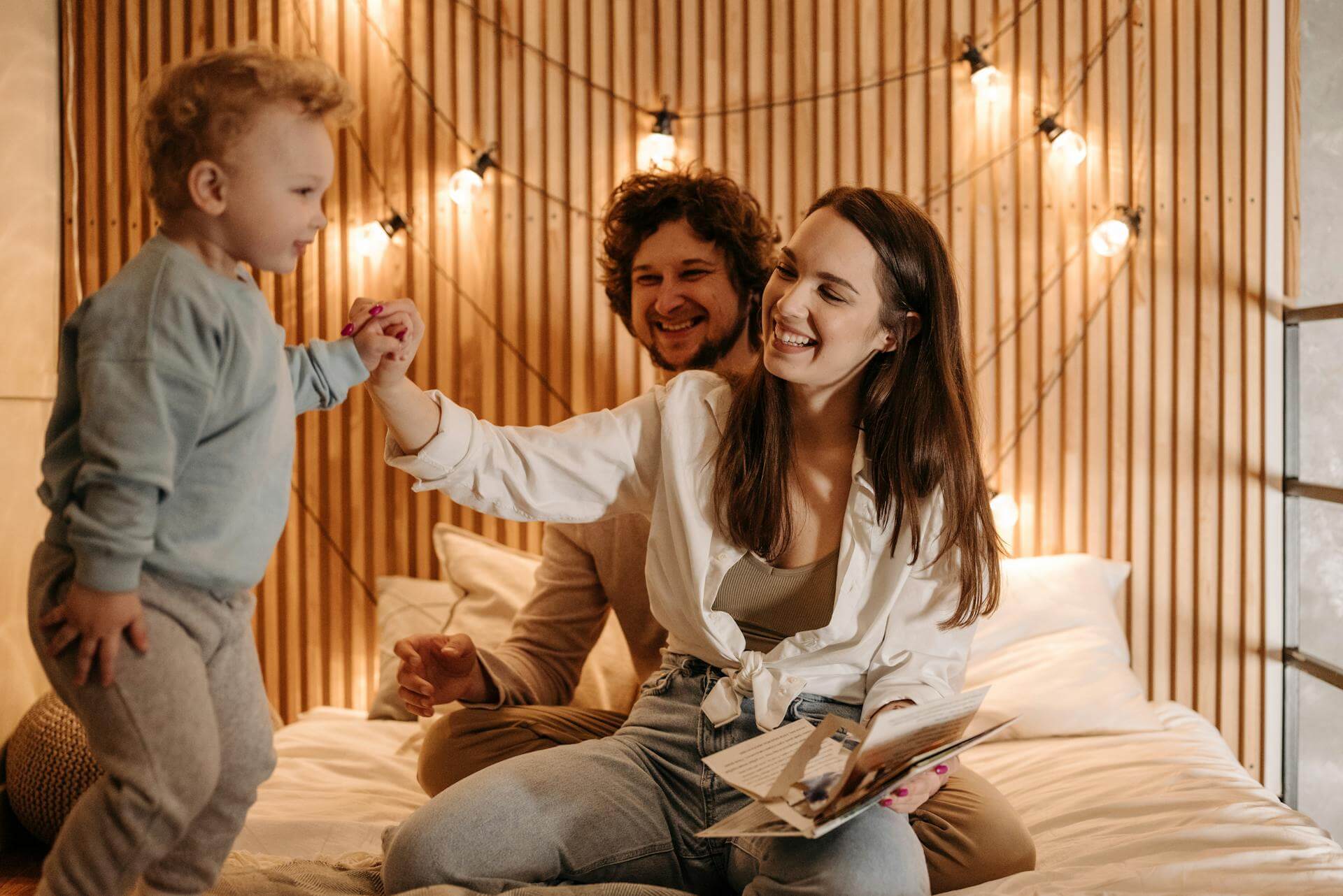 A man and woman sit on a bed, smiling at each other while a child plays happily between them