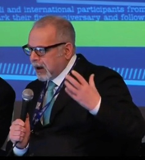 A photo of a man wearing glasses and a suit giving a lecture while sitting in front of a blue and green screen.