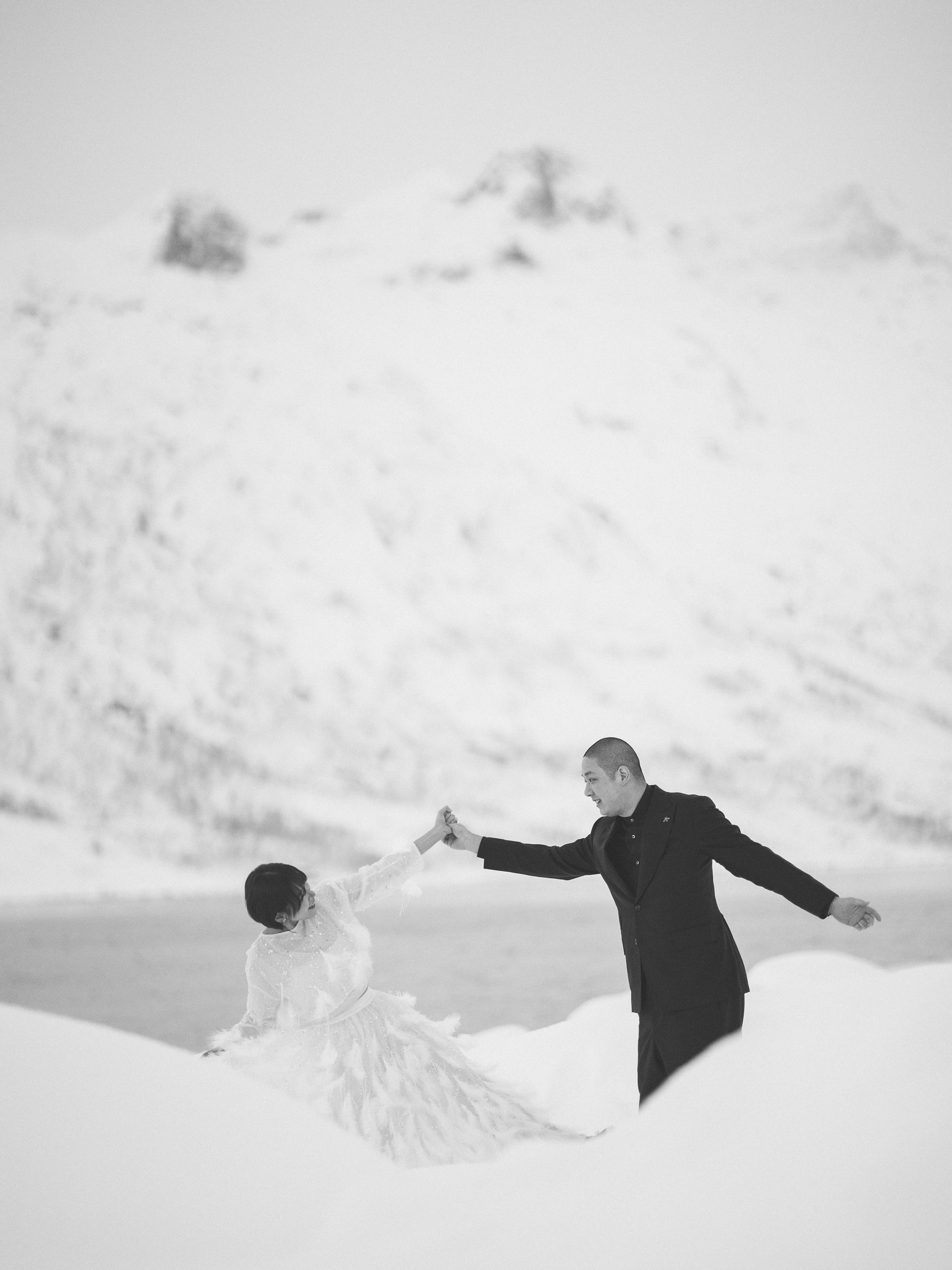 A distant, monochrome view shows the couple reaching toward one another across a broad, nearly abstract snow field.