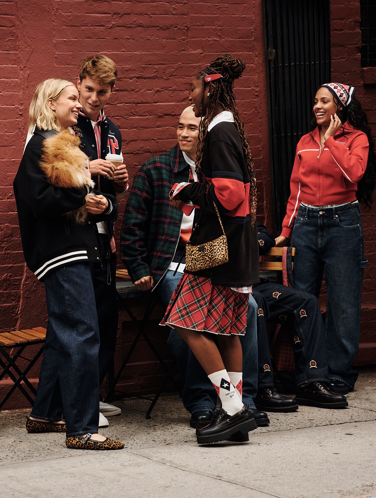 A group of young people chat and laugh on a city sidewalk, wearing casual Tommy Hilfiger outfits against a red brick wall.