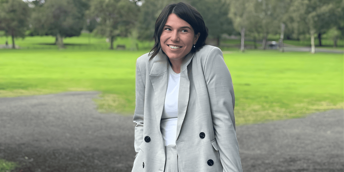 Smiling woman in light gray blazer standing on park path with green lawn and trees behind.