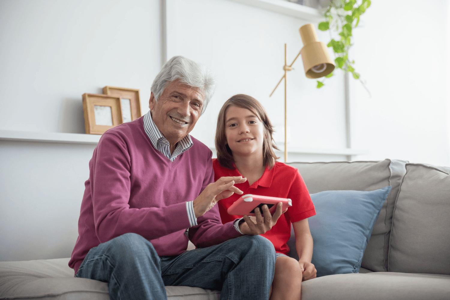An older man and his grandson side by side on the sofa with a tablet held between them, both smiling.