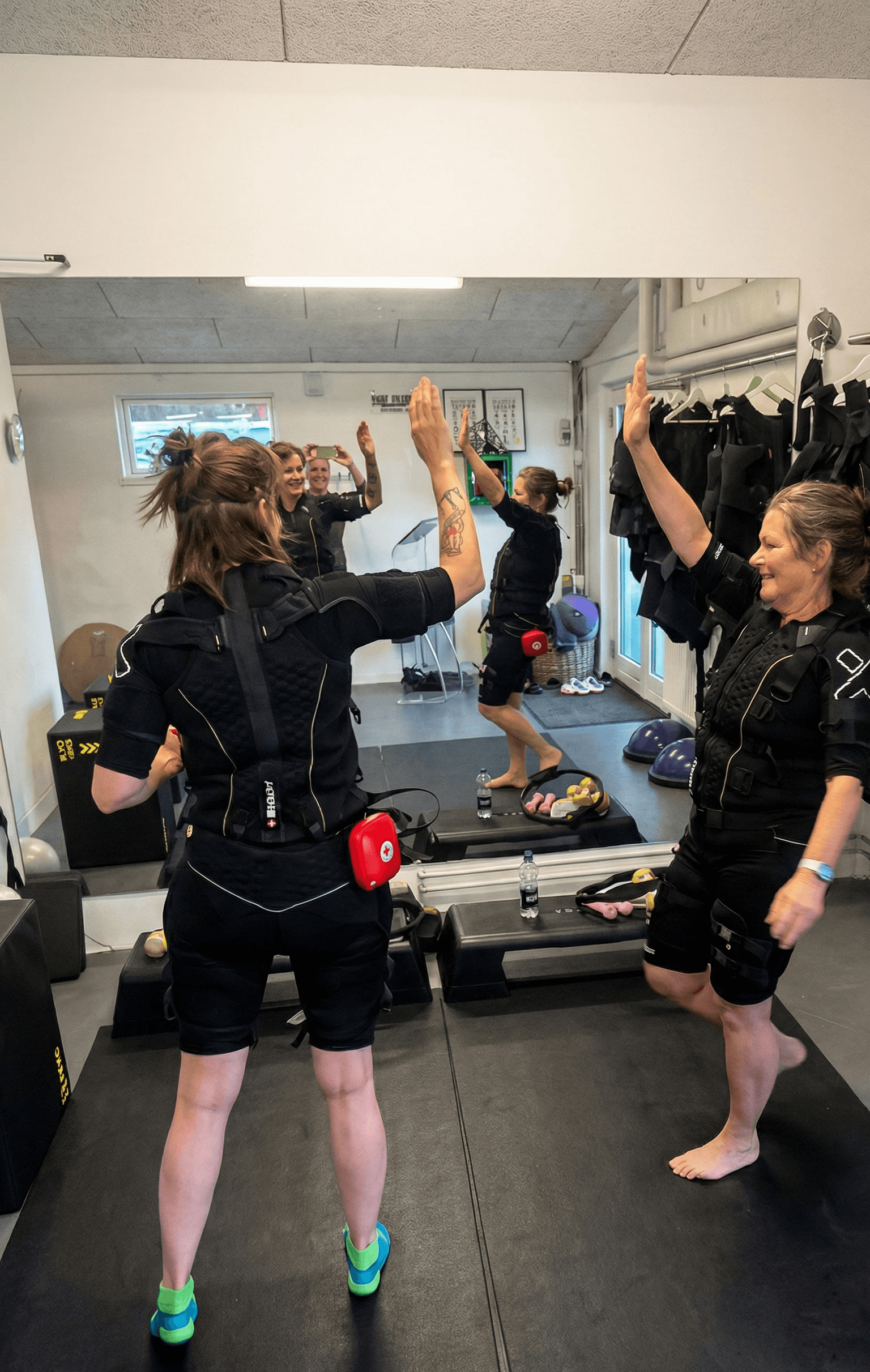 A dimly lit gym scene featuring a barbell rack and weights on the floor, with large windows in the background.