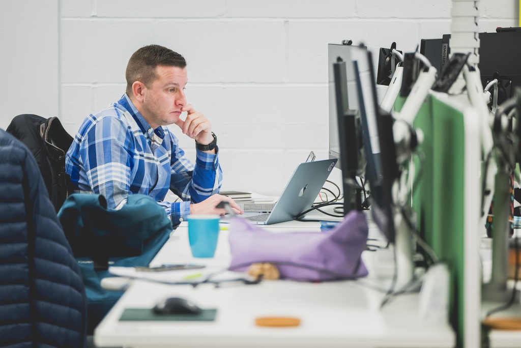 An open-plan office environment with a person seated at a long desk working on a laptop connected to multiple desktop monitors. The workspace includes a keyboard, mouse, notebooks, and a blue reusable cup, with coats draped over nearby chairs. The setting features a white brick wall, green privacy dividers between desks, and soft, natural lighting that gives the space a casual, collaborative feel.