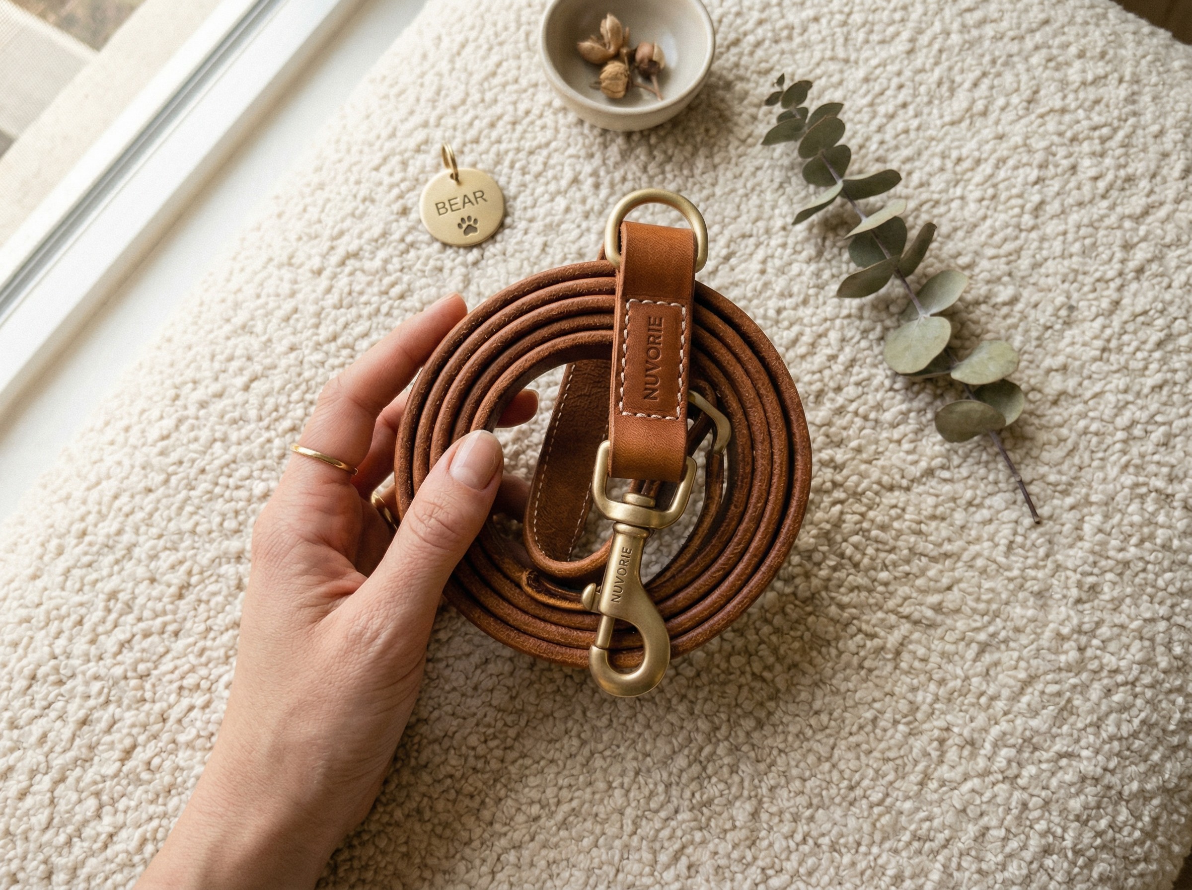 A person's hand holding a rolled Nuvorie premium cognac leather dog leash with brass hardware on a textured boucle fabric
