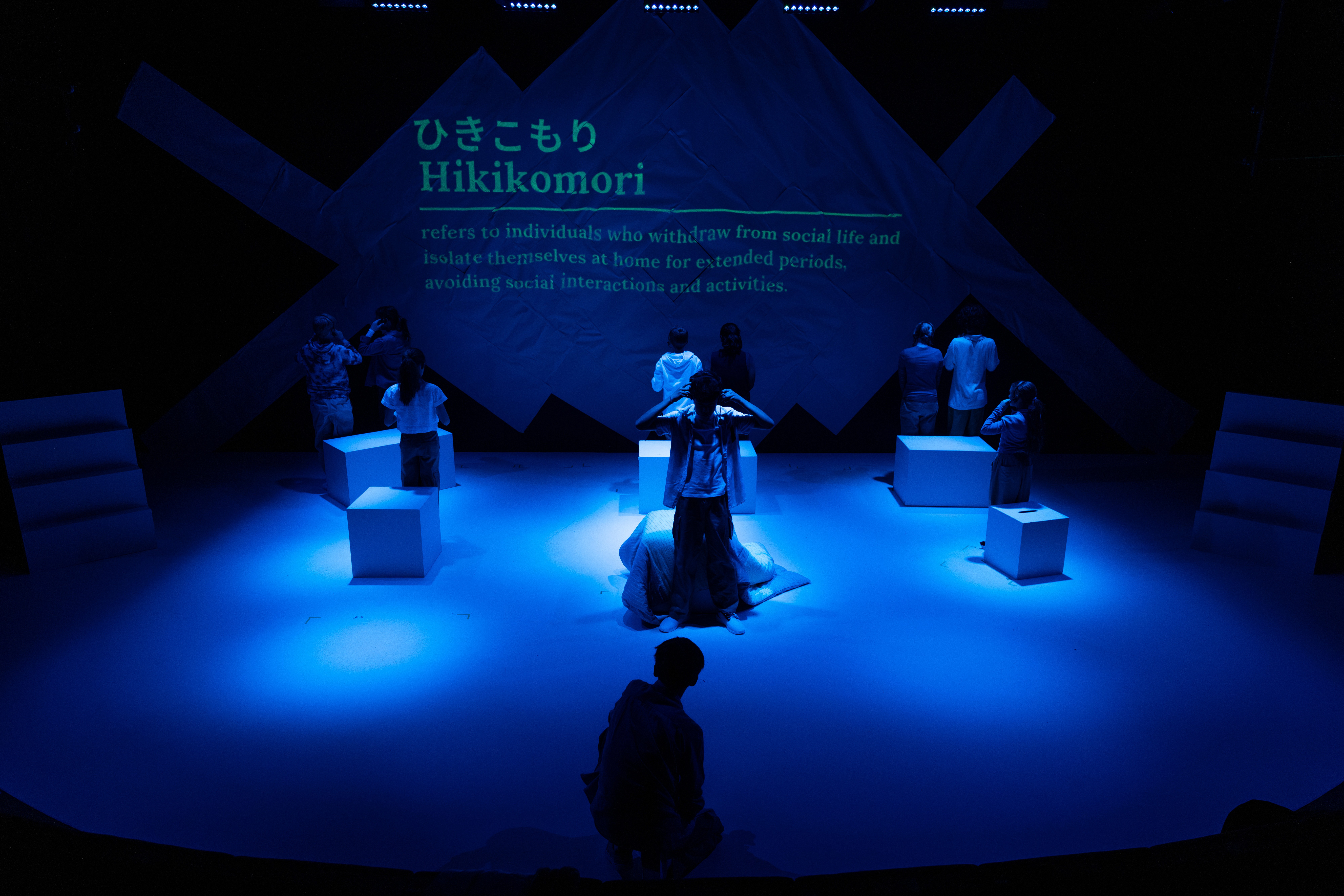 With the stage washed in deep blue light, performers stand facing away from the audience as projected text defining “Hikikomori” appears on a textured paper-like backdrop.