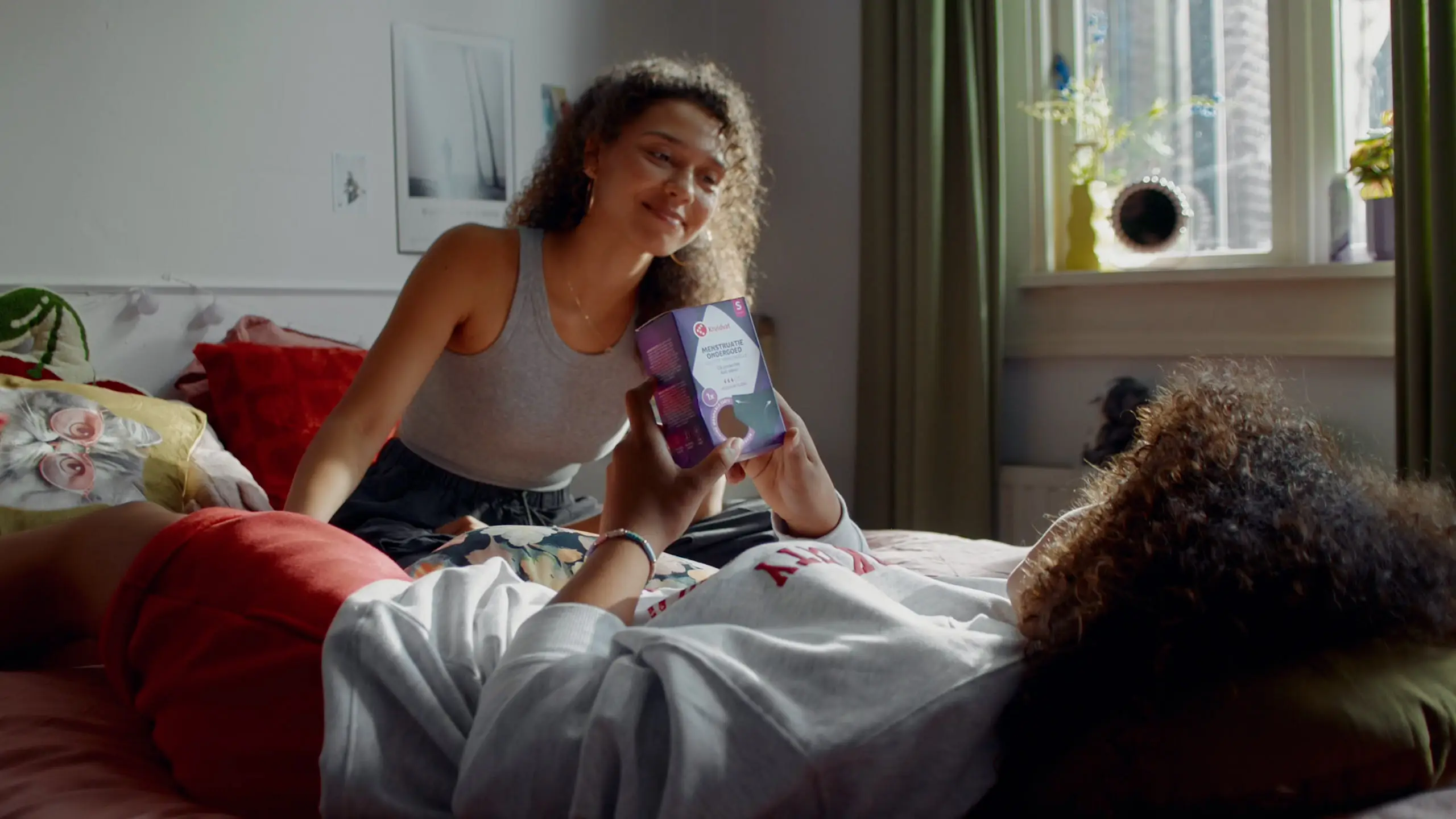 Two sisters on a bed, one holding up a Kruidvat menstrual underwear box to show the other.