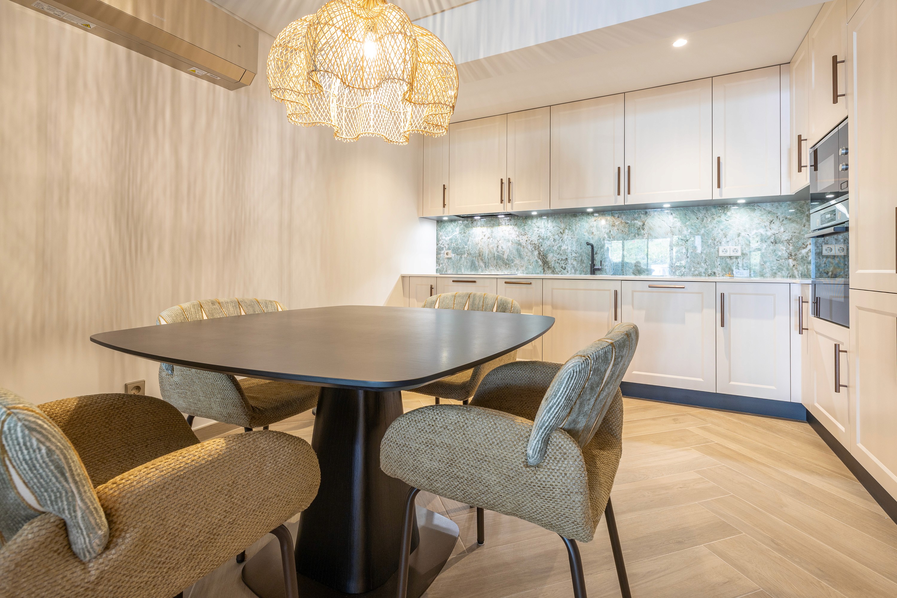 Open kitchen in pale oak against green veined stone, with a round dining table and a wicker pendant