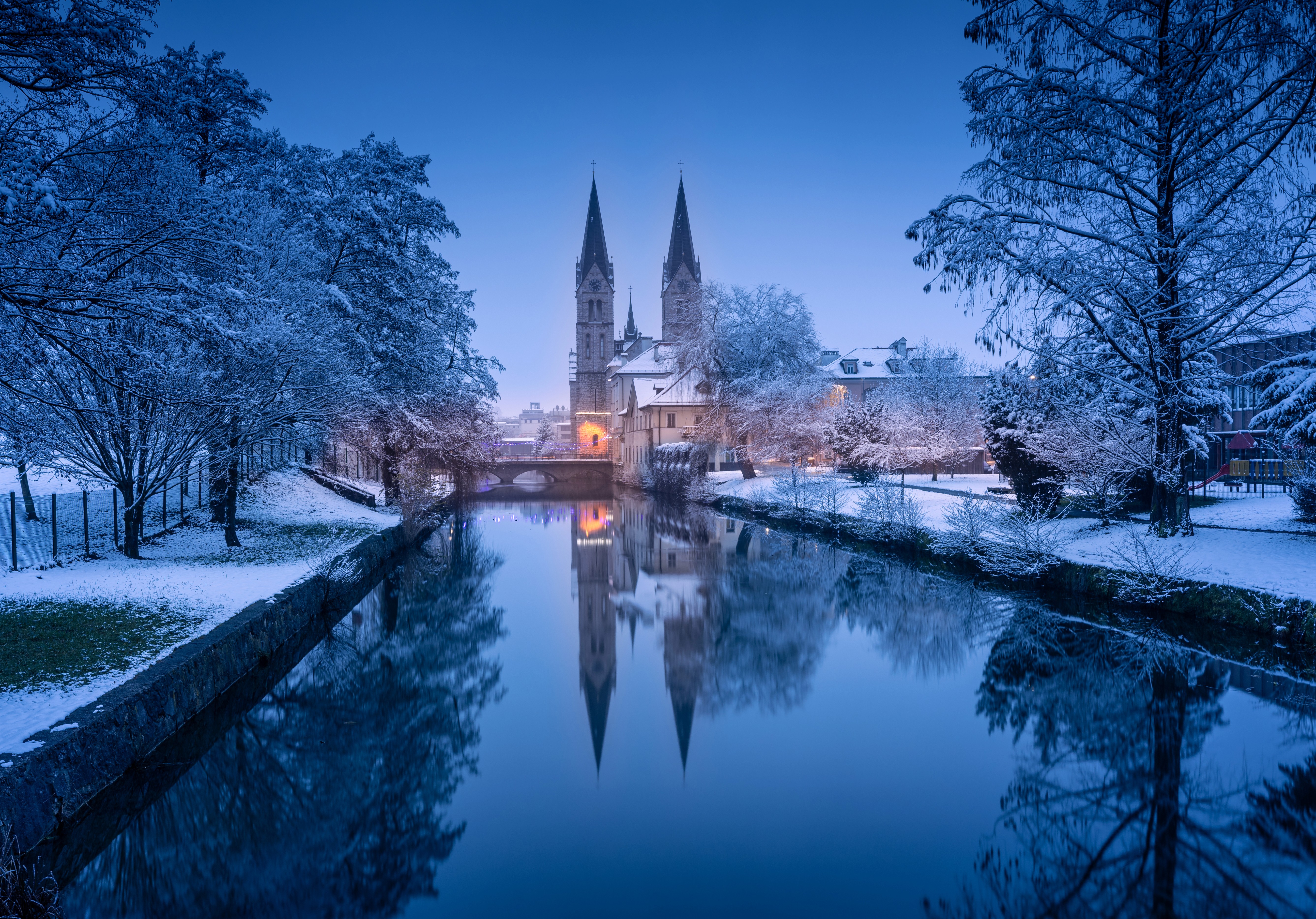 The neo-Romanesque Church of St. Bartholomew in Kočevje, Slovenia, reflected in the calm Rinža River during a blue hour winter evening with snow-covered trees and a small bridge.