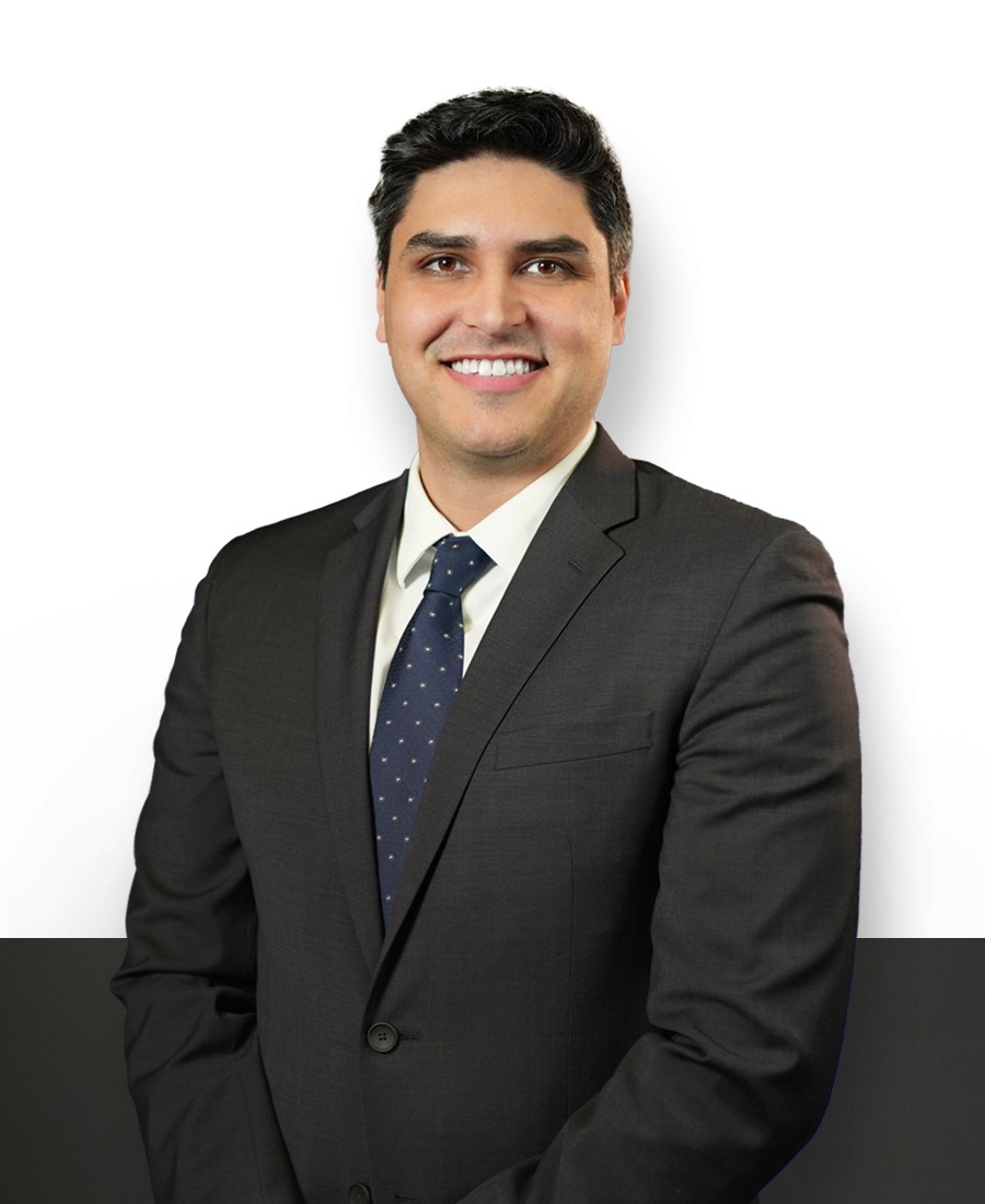 corporate headshot of a male with warm light brown skin and black hair, smiling broadly in a black suit with a white shirt and navy patterned tie. 