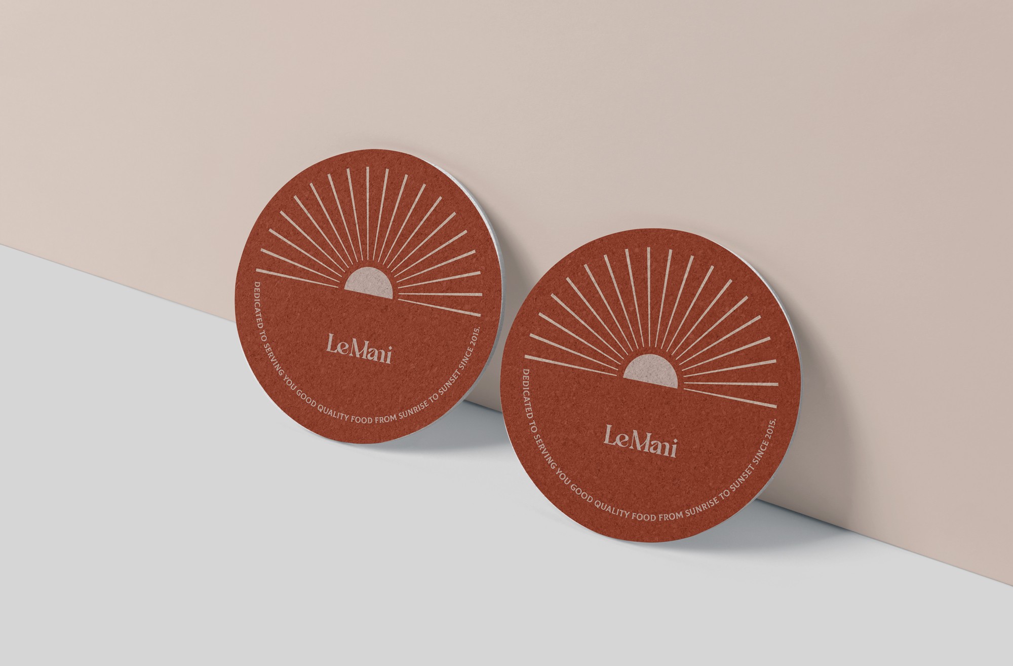 這張圖片展示了兩個圓形的Le Mani杯墊,顏色為深紅色,上面印有簡約的太陽圖案和品牌名稱。