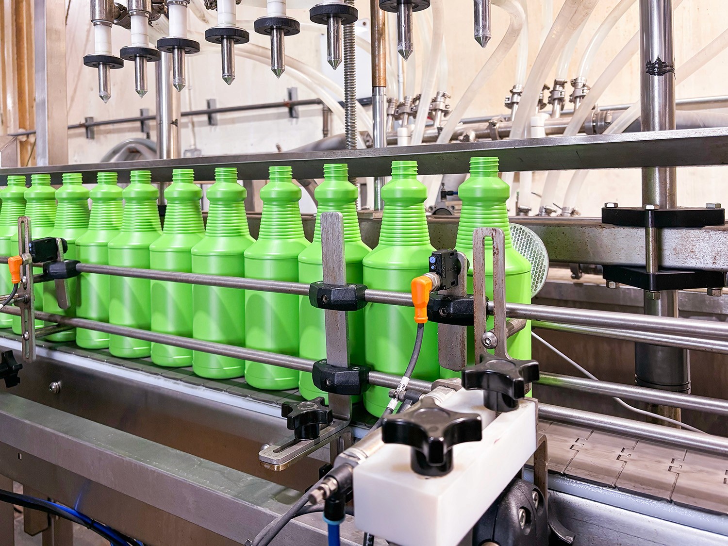 Green plastic bottles on a conveyor belt in a chemical manufacturing facility, being filled by automated machinery.