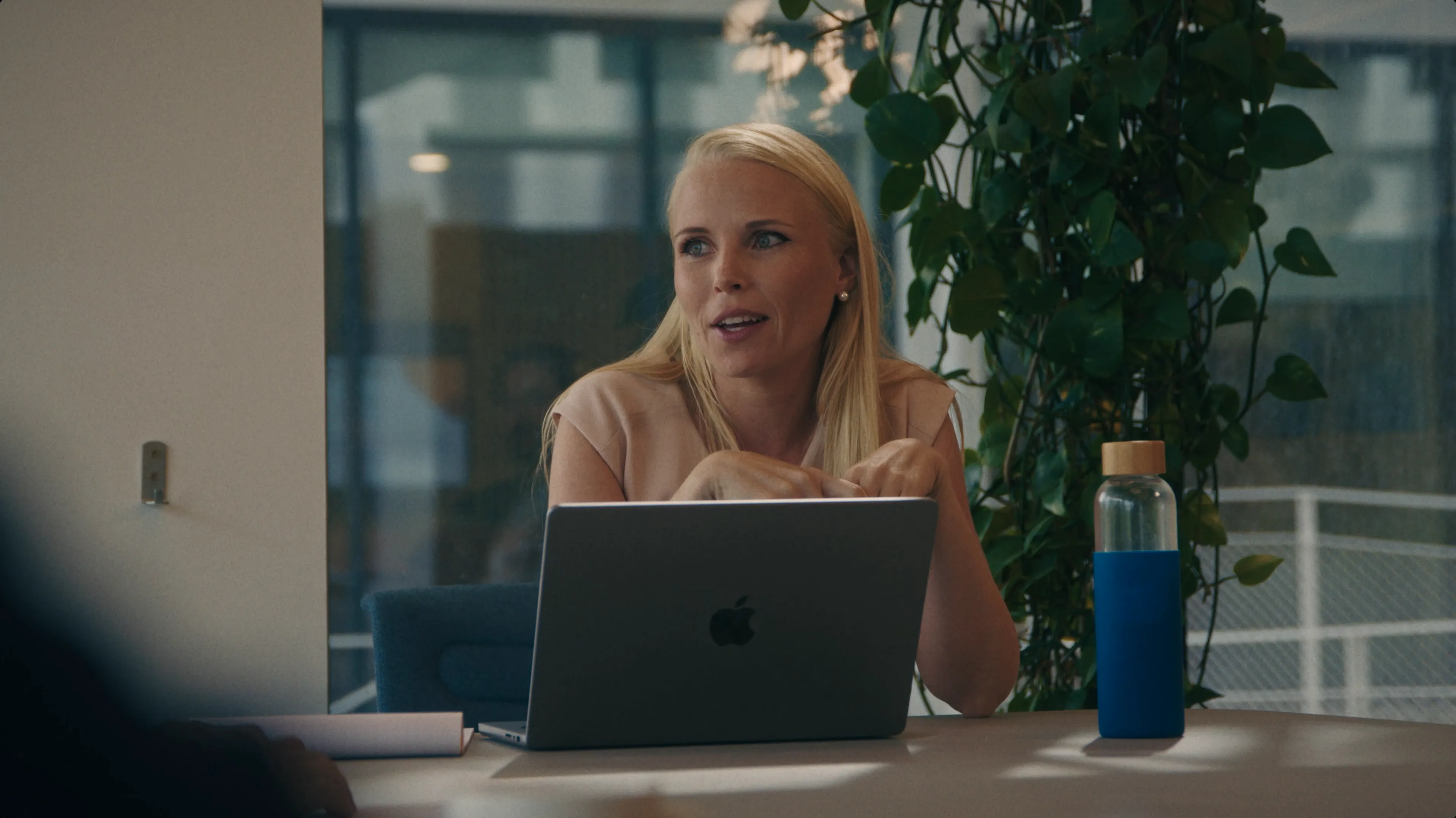 A woman with blonde hair sits at a table in an office, engaging in conversation, with an open laptop, a notebook, and a reusable water bottle nearby, while green plants add a touch of nature to the modern workspace setting.