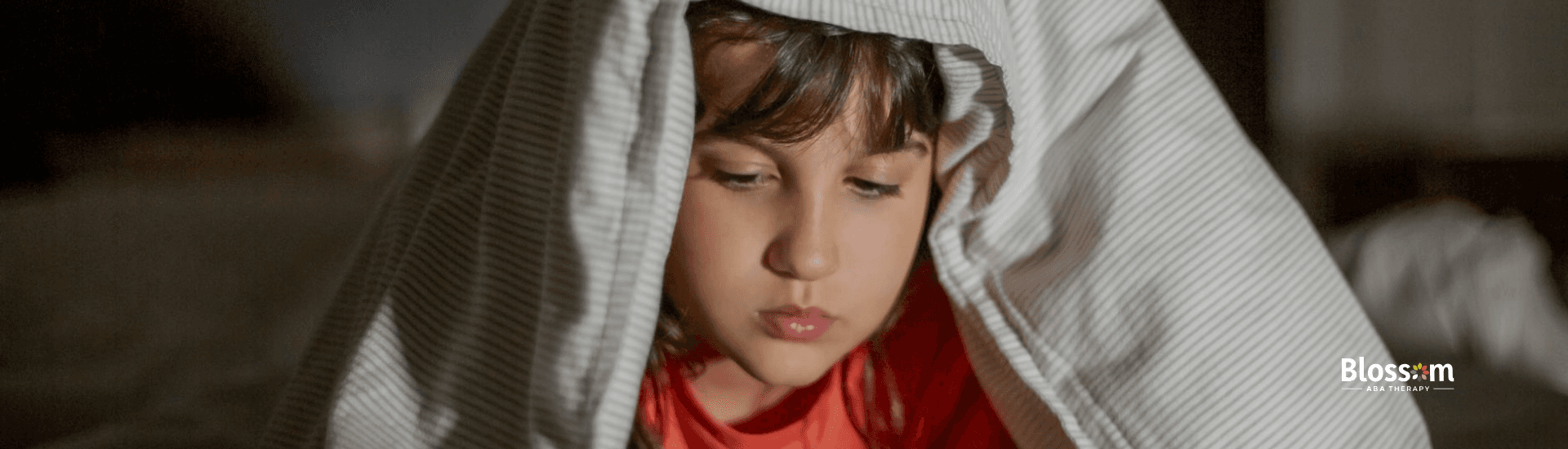 An autistic girl under a blanket in a dimly lit room seriously watches a tablet screen in Georgia.