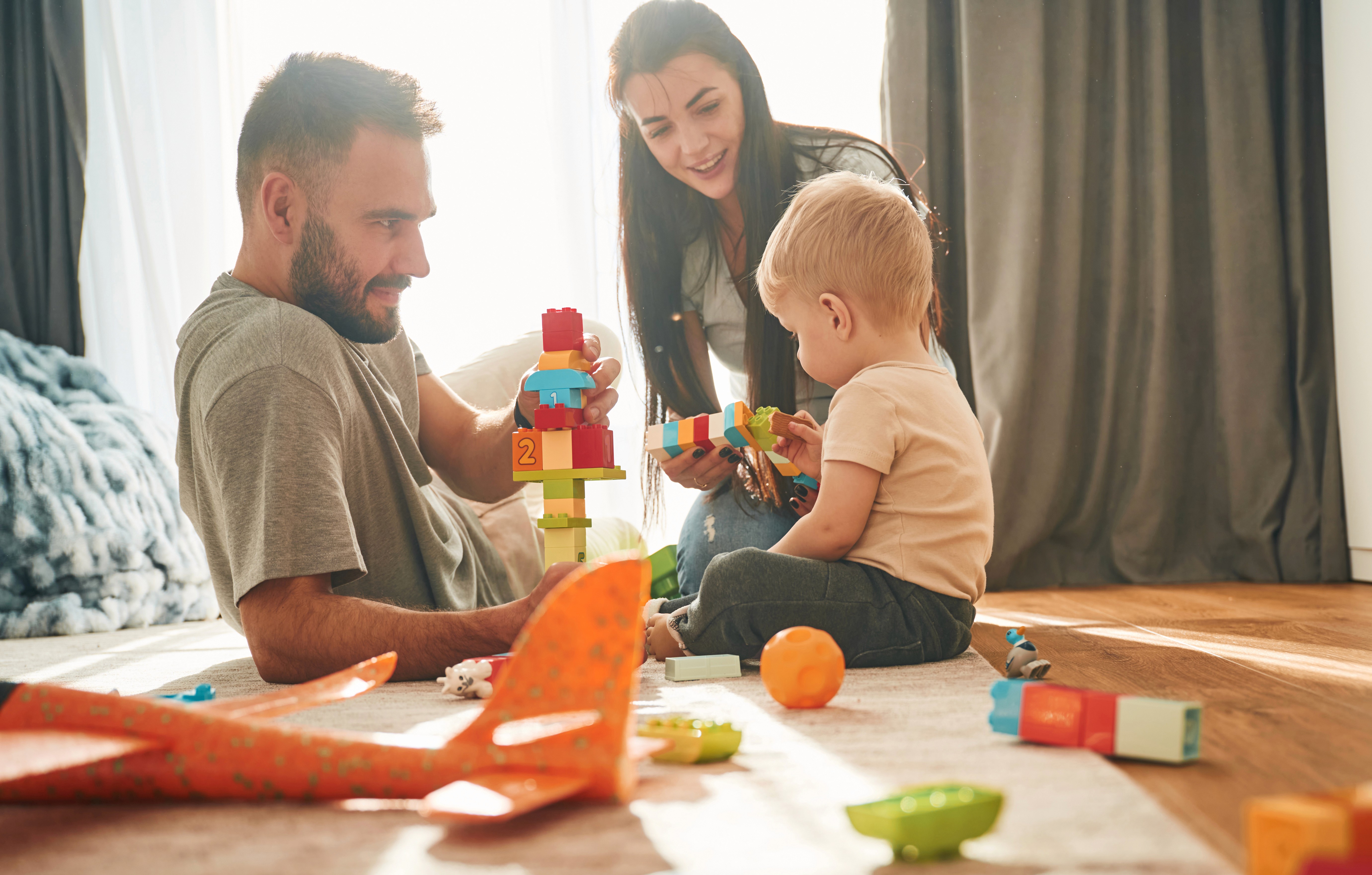 Eltern spielen gemeinsam mit ihrem Kleinkind auf dem Boden mit Bauklötzen im Wohnzimmer.