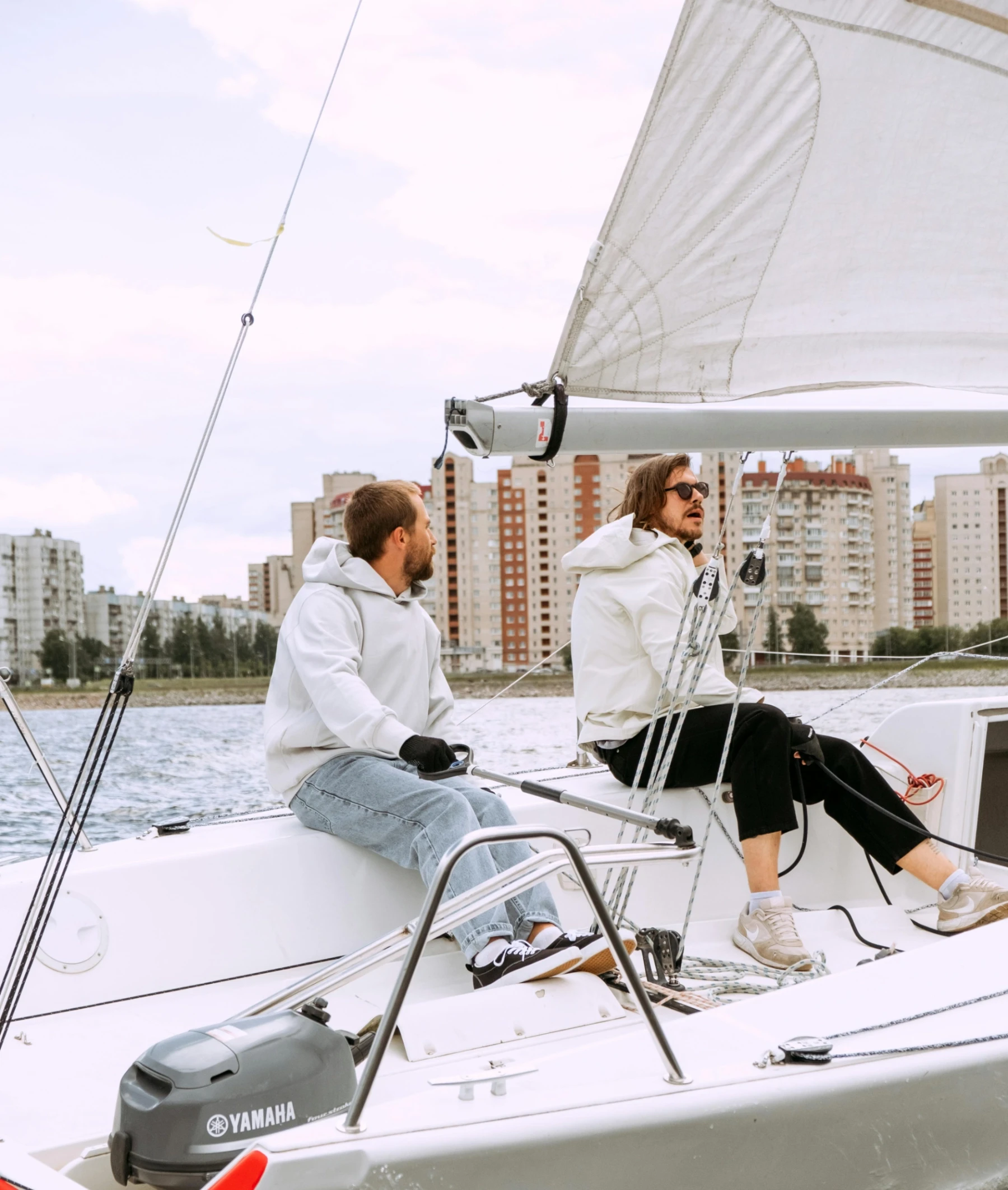 Two people in casual attire sail a small boat on a city waterfront. They focus on steering, with modern buildings in the background and a relaxed atmosphere.