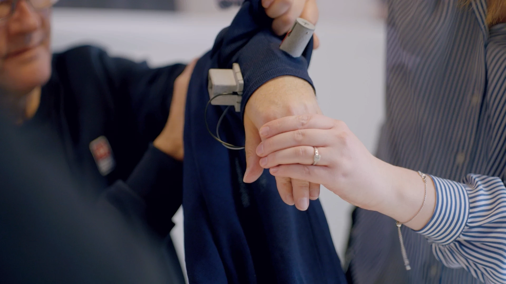 Close-up of an older person's arm fitted with a wearable monitoring device while two people adjust it.