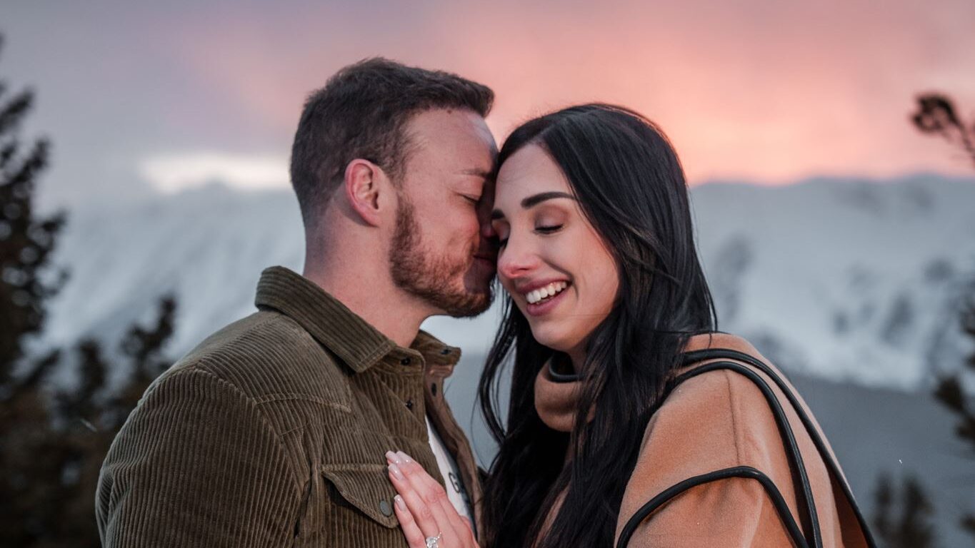 Newly engaged couple close together during alpenglow at Smith Cabin Aspen, pink and purple sky over snow-covered mountains behind them, engagement ring visible