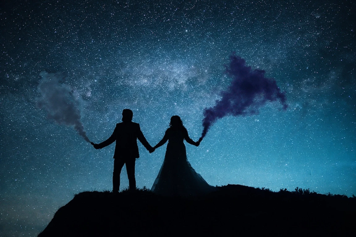 Silhouette of bride and groom holding smoke bombs beneath a star-filled night sky during a creative wedding portrait