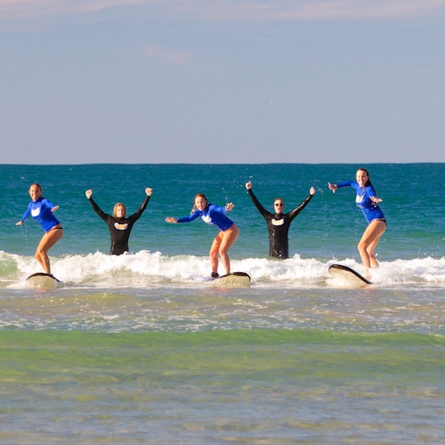 Limang tao na naka-wetsuit ang nagse-surf sa mababaw na alon ng dagat, itinaas ang kanilang mga kamay sa tila masiglang pagbati.