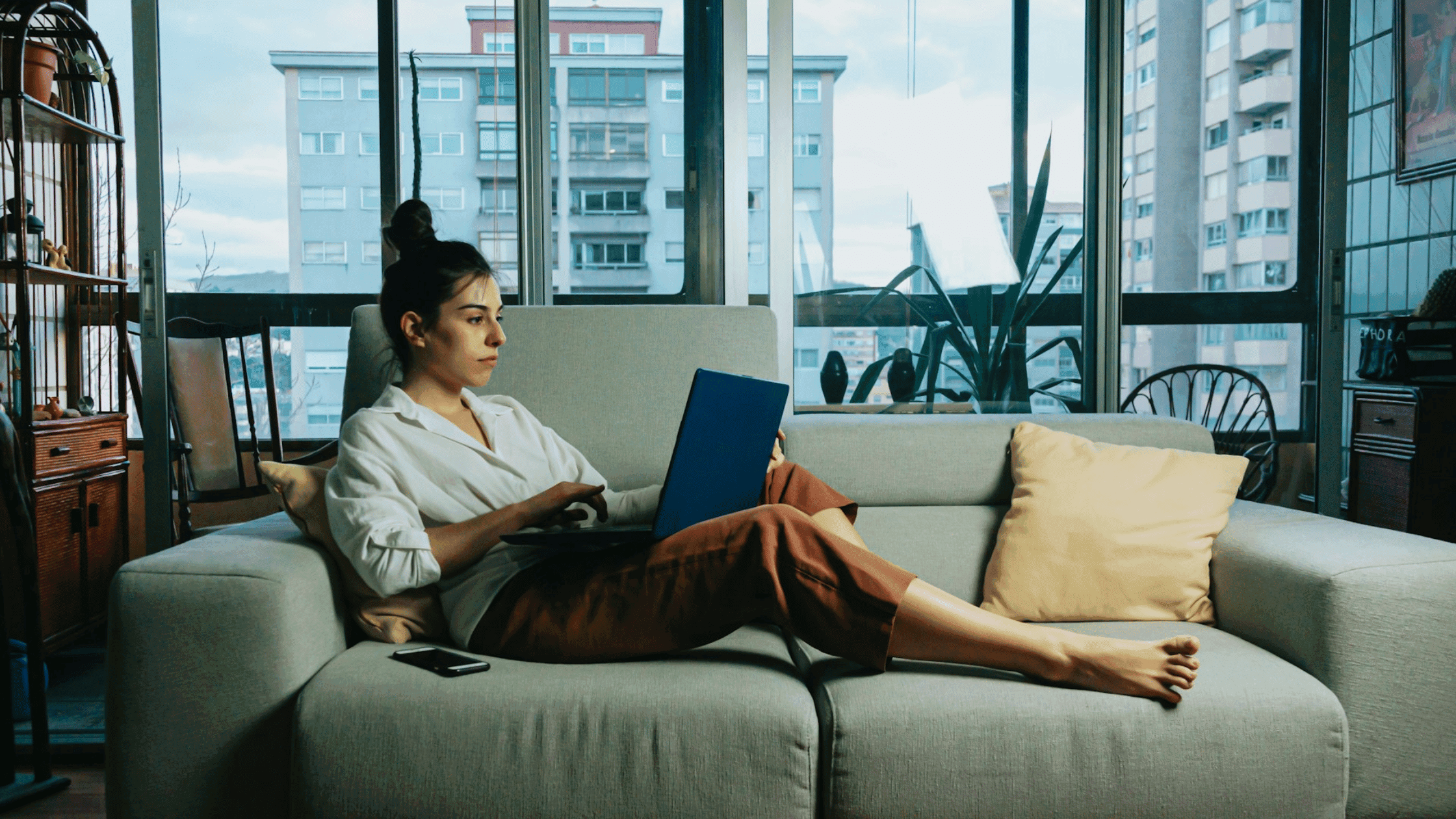 The image shows a young woman intently using a laptop while seated on a couch in an office-like setting with a cityscape visible through the windows.