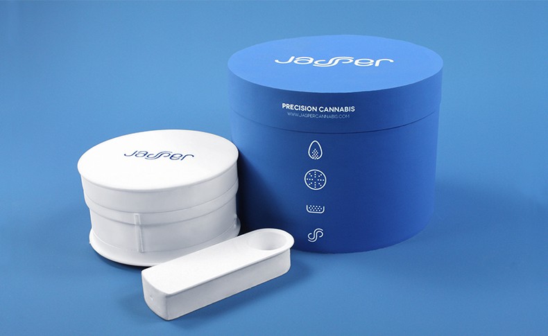 A blue cylindrical container labeled "Jasper" and "Precision Cannabis" sits on a blue surface next to a white lid and a white tube. The container has three small icons on its side: a leaf, a circle, and a wavy line. The lid also displays "Jasper" in white text. The tube is positioned diagonally in front of the container.