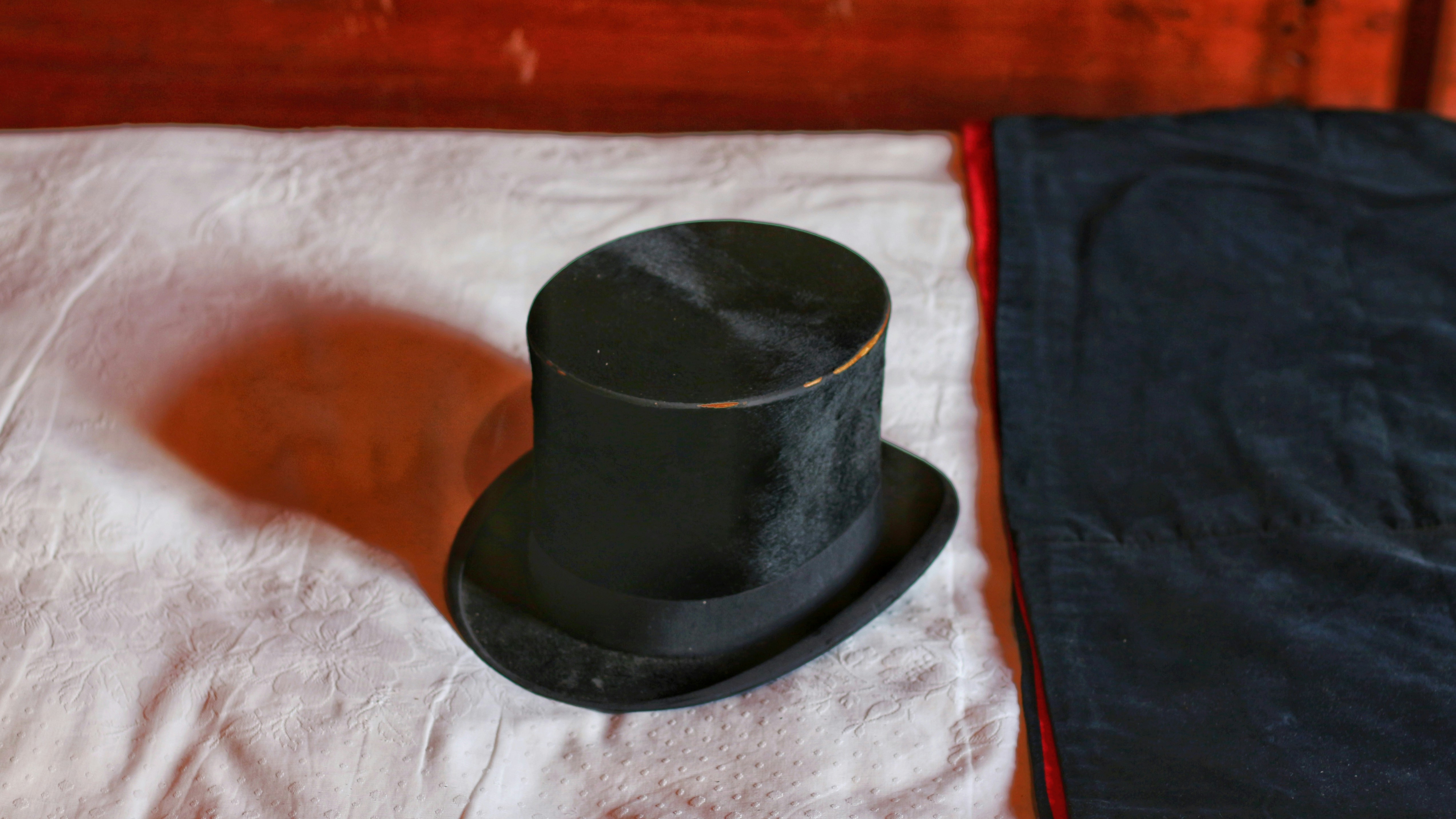 A black top hat placed on a white fabric surface beside folded dark clothing.