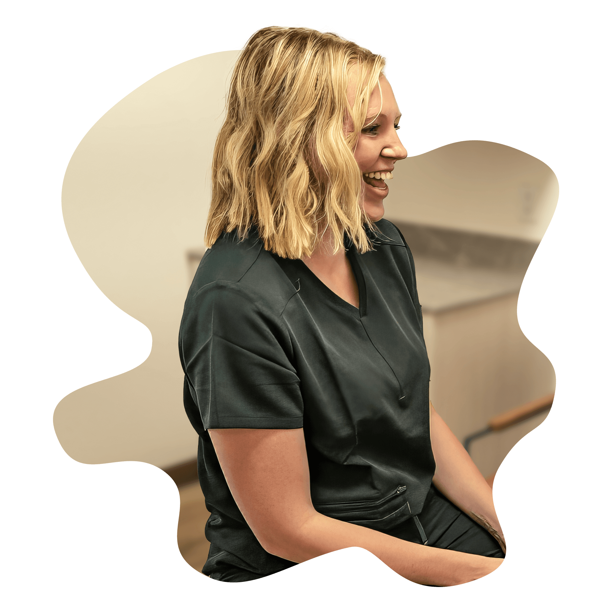 A woman with blonde hair smiles while seated, wearing a dark shirt against a neutral background