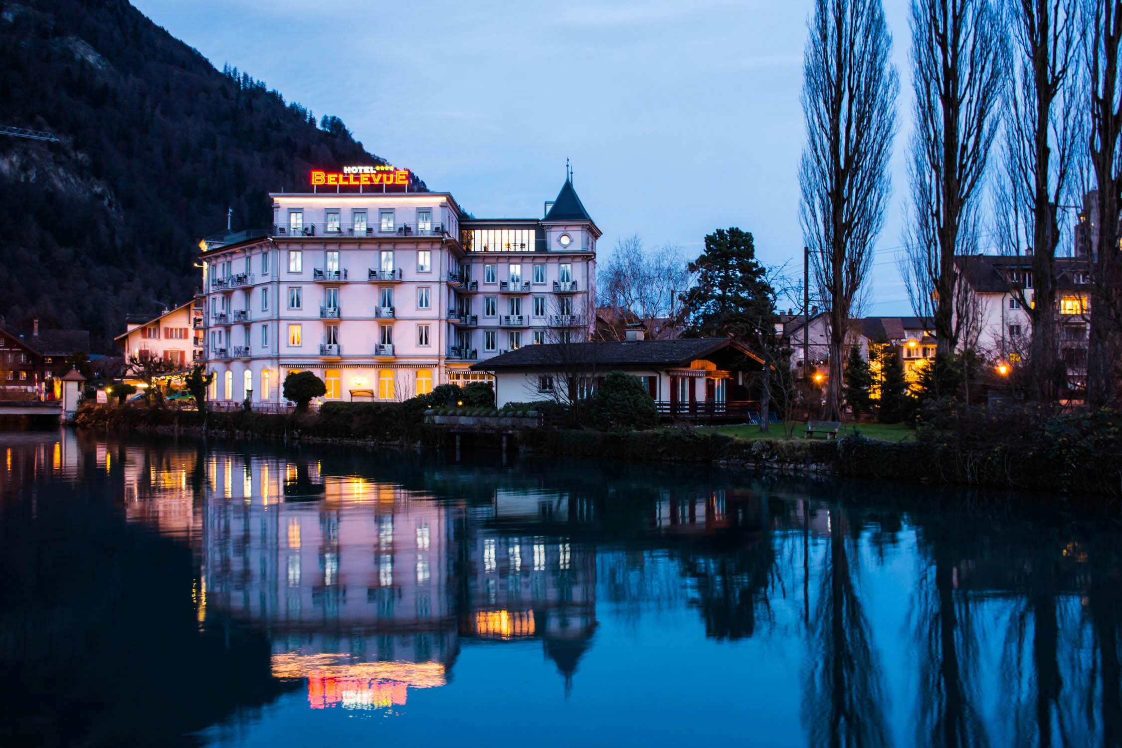 A large white hotel with a red sign sits on a lakefront at dusk, reflected in the water beneath tall poplar trees.