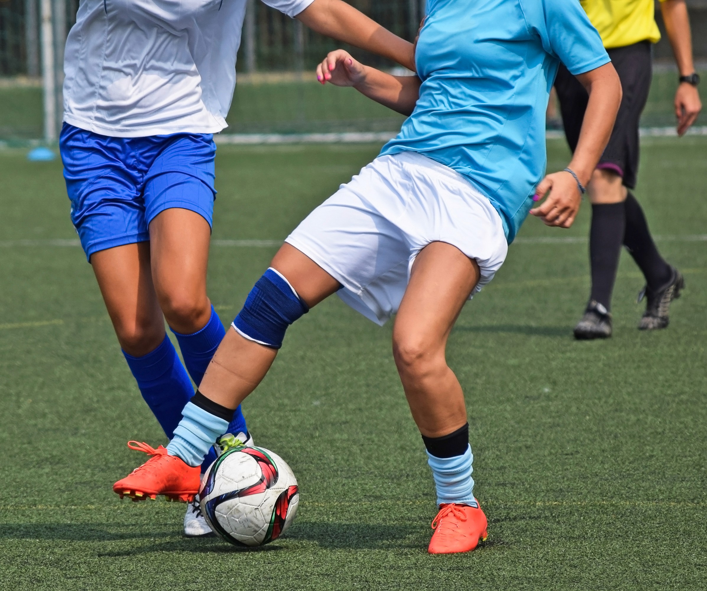 Imagem de duas jogadoras de futebol em ação durante uma partida. Ambas estão disputando a posse de bola, com uma delas usando chuteiras laranja e a outra chuteiras brancas. A jogadora à esquerda veste shorts e meias azuis e a camisa branca, enquanto a jogadora à direita usa camisa azul claro e shorts brancos com uma joelheira azul. O campo é de grama sintética e um árbitro pode ser visto ao fundo. A cena captura a intensidade e competitividade do esporte.