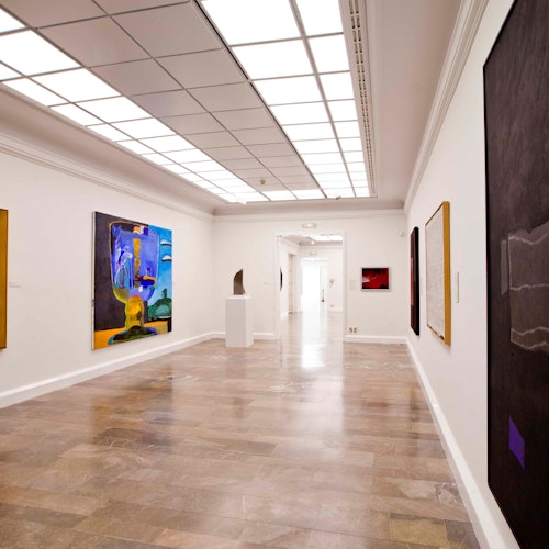 一個藝術畫廊的走廊,白色牆壁上掛滿色彩斑斕的抽象畫, pedestal 上有一座雕塑,地板是瓷磚的。