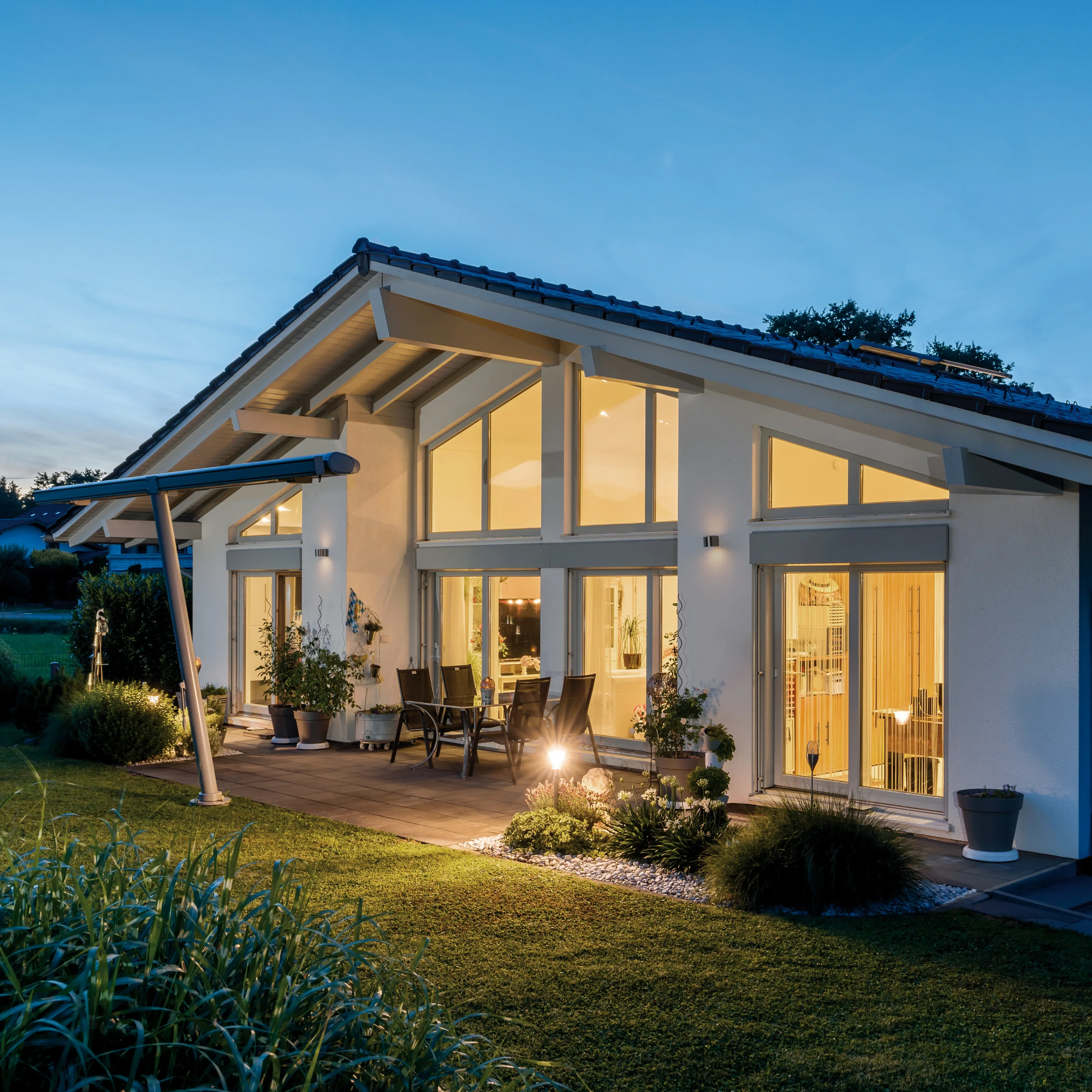 Modernes Einfamilienhaus mit großen Fenstern und beleuchteter Terrasse im Abendlicht, Garten mit gepflegtem Rasen.