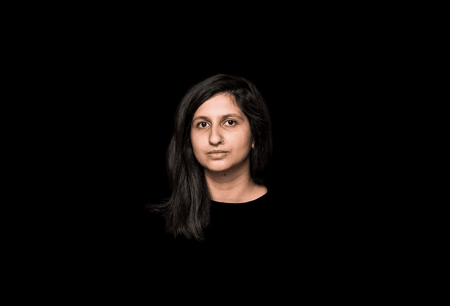 A woman with long, dark hair is shown against a solid black background. She is wearing a dark top and has a neutral expression. Her face is centered in the frame.