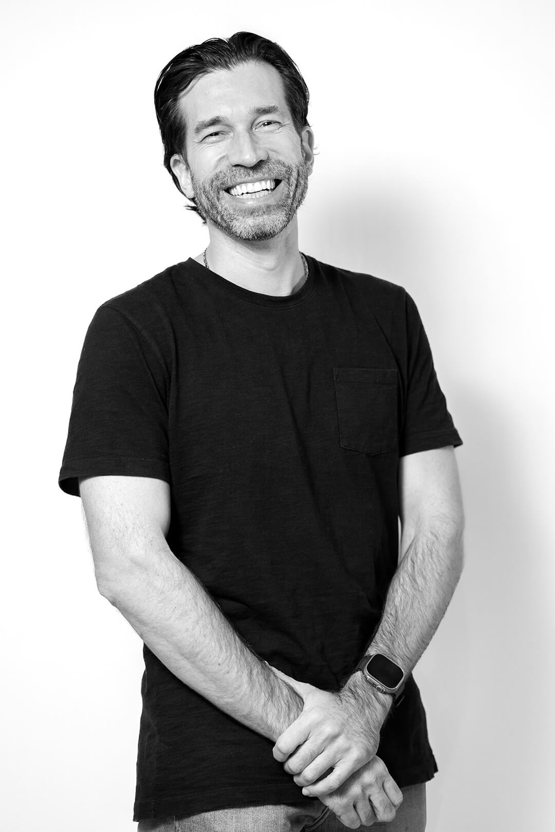 Smiling man in a black t-shirt stands against a white background. His hands are clasped in front, exuding a relaxed and cheerful demeanor.
