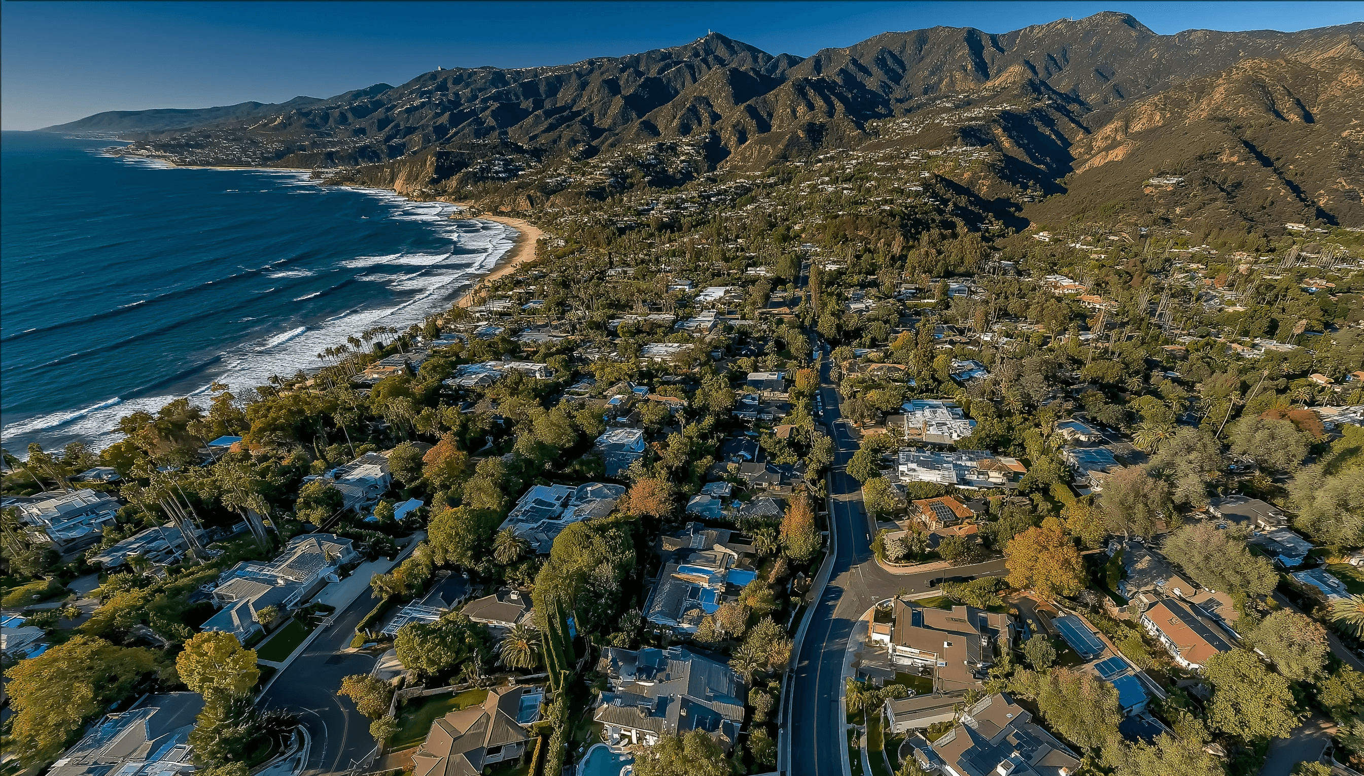 Aerial view of an affluent coastal neighborhood with lush green trees and luxury homes nestled between the bright blue ocean and rugged mountains under a clear sky.
