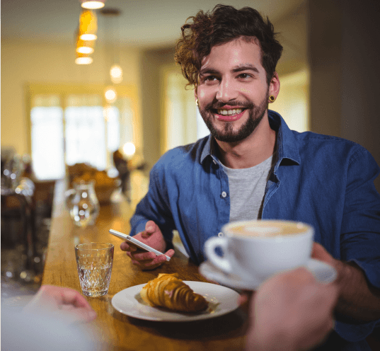 Un collage de cuatro fotos que muestran un café. En una imagen, una persona está usando un teléfono móvil que muestra un descuento del 50% por Black Friday. Otra muestra a una mujer detrás del mostrador con una tableta. La tercera imagen retrata el interior de un café moderno y elegante, con bicicletas apoyadas contra la pared, y la cuarta muestra a un hombre absorto en su teléfono. El café se llama "Morrow Cafe" y ofrece un cashback del 12%.