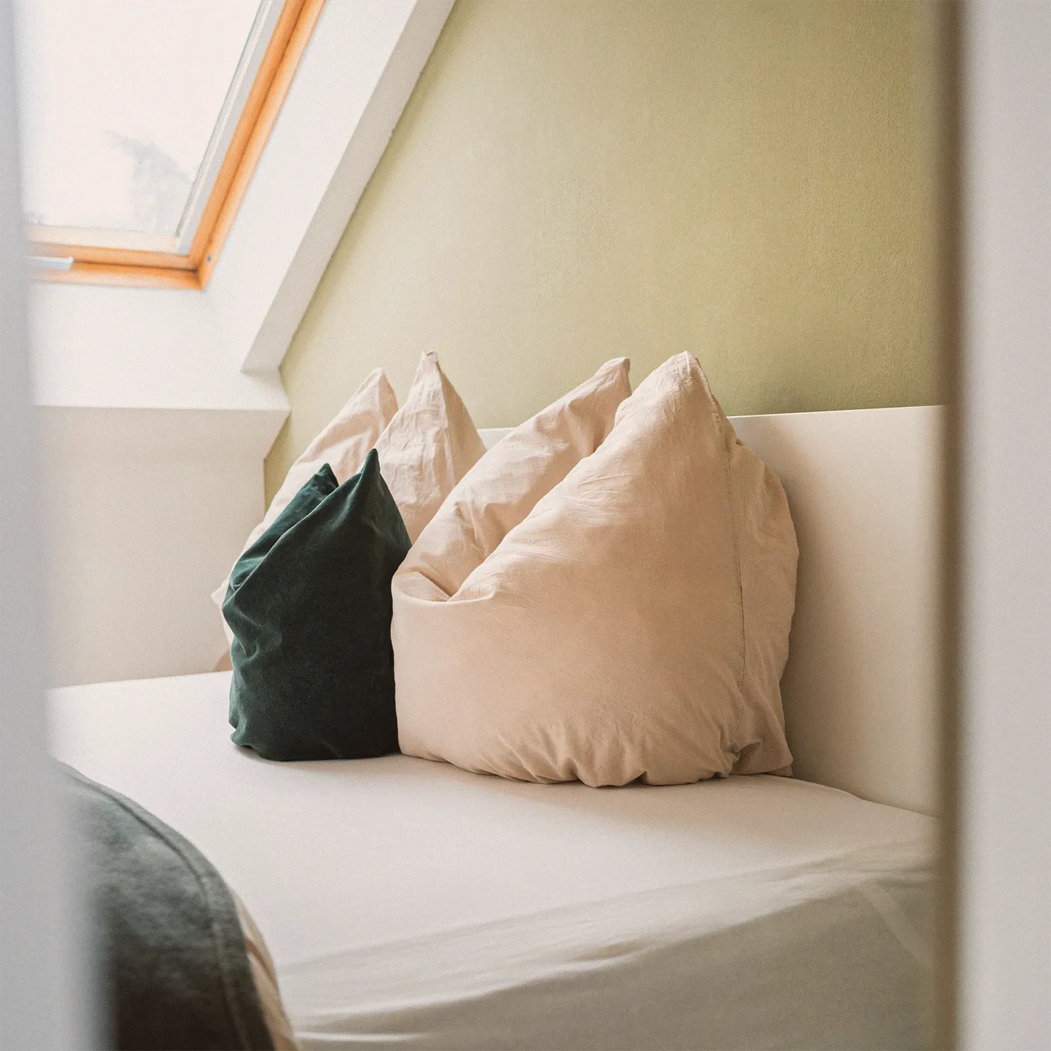 Loft N°1 • bed with linen and green velvet cushions under a skylight and an olive-green wall