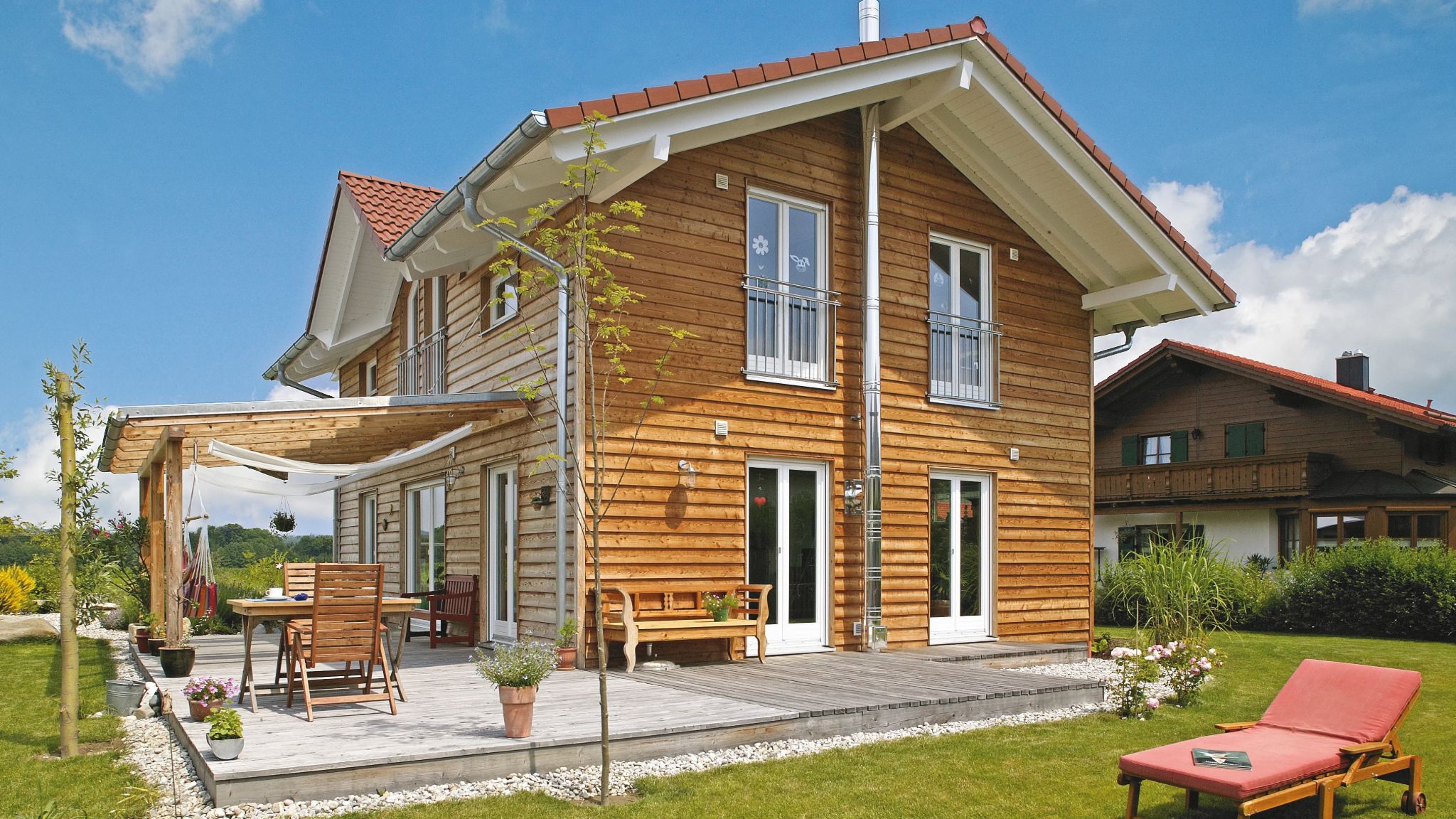 Holzhaus mit Terrasse, Gartenmöbeln, Hängematte und Liege unter blauem Himmel.