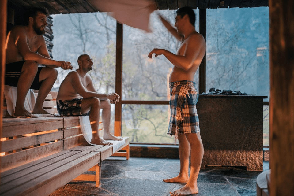 Three men relax in a warm, wood-paneled sauna, with one man wearing a towel around his waist and standing, while the others sit on a bench, bathed in soft, ambient light from a large window.