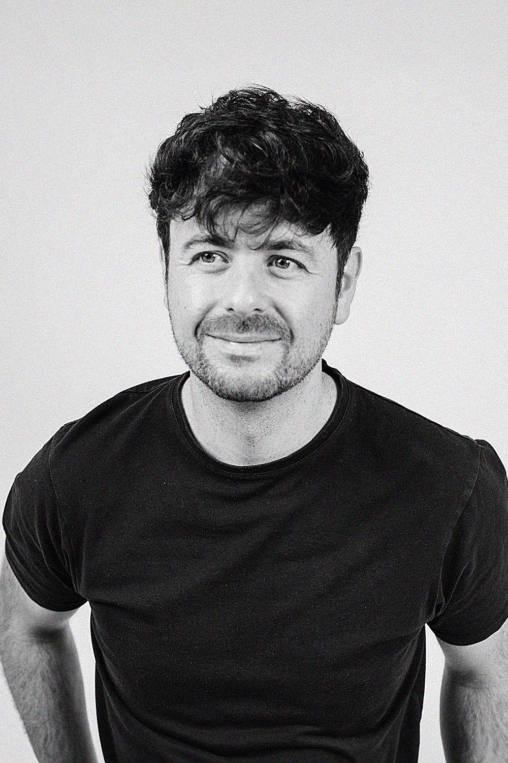 A smiling person with curly hair, wearing a leather jacket, stands in a cozy indoor setting.