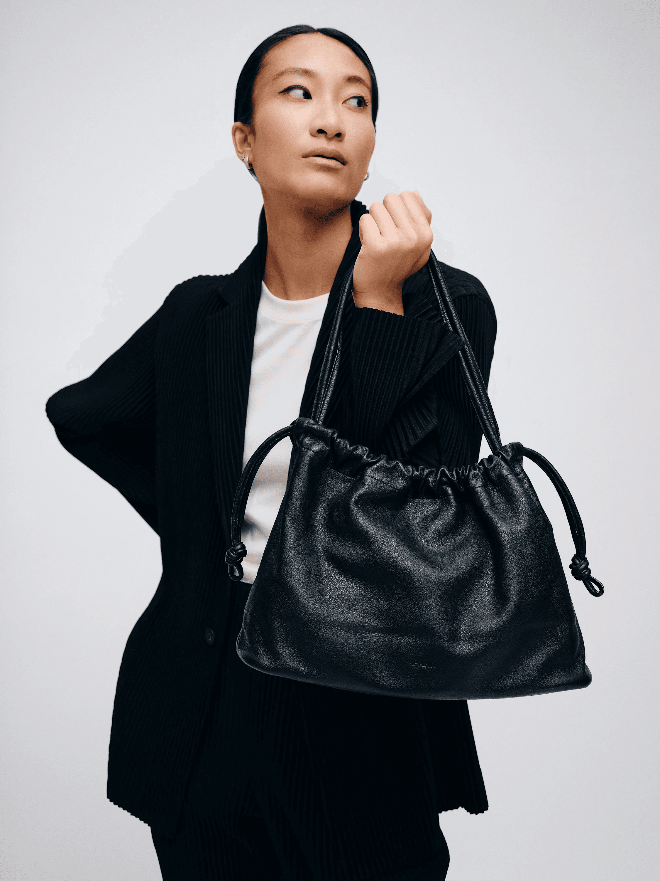 A woman in a black blazer holds a stylish black leather handbag with a drawstring closure, set against a neutral background.