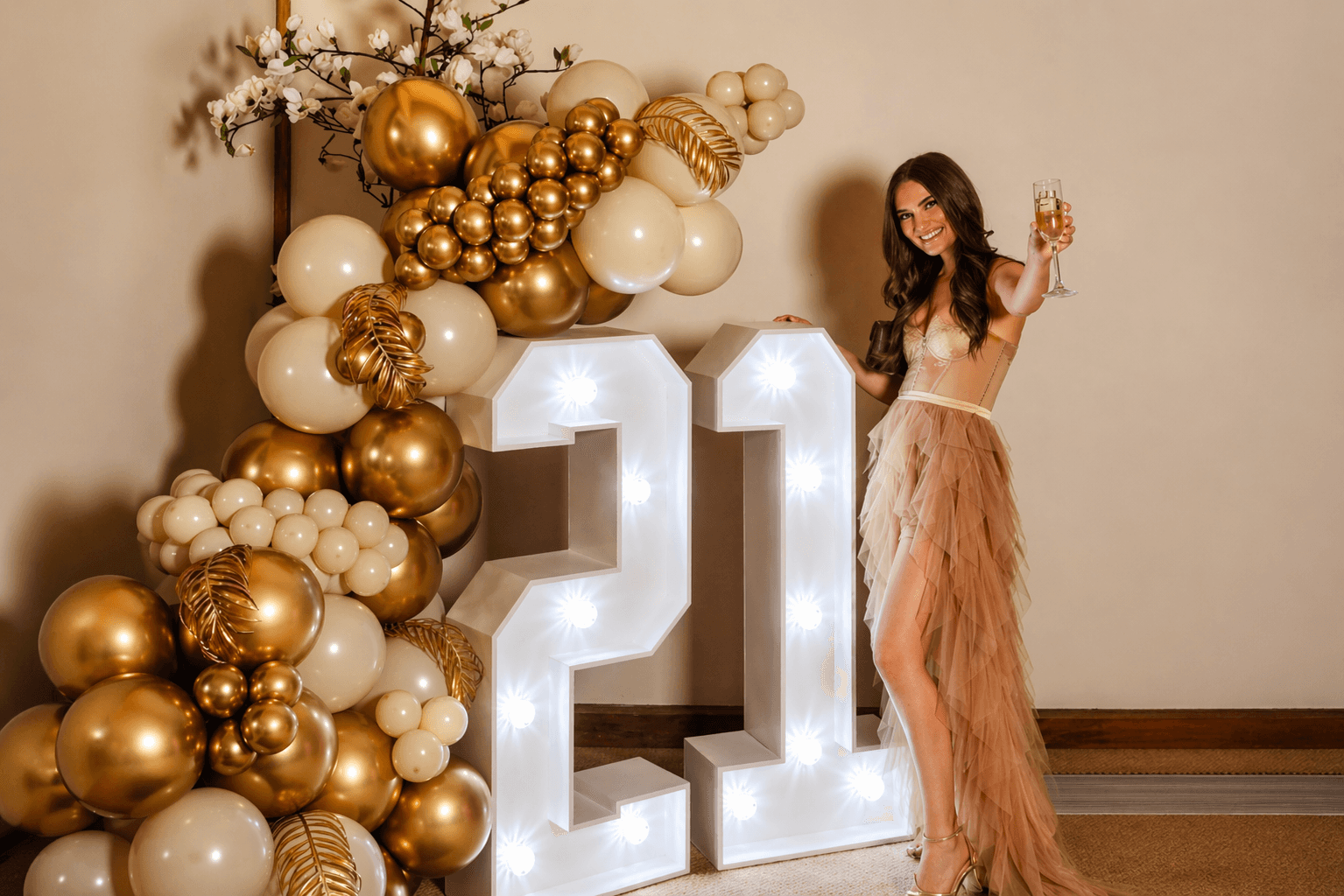 Brunette woman raises champagne glass next to lit "21" sign with gold/white balloons & flowers in formal