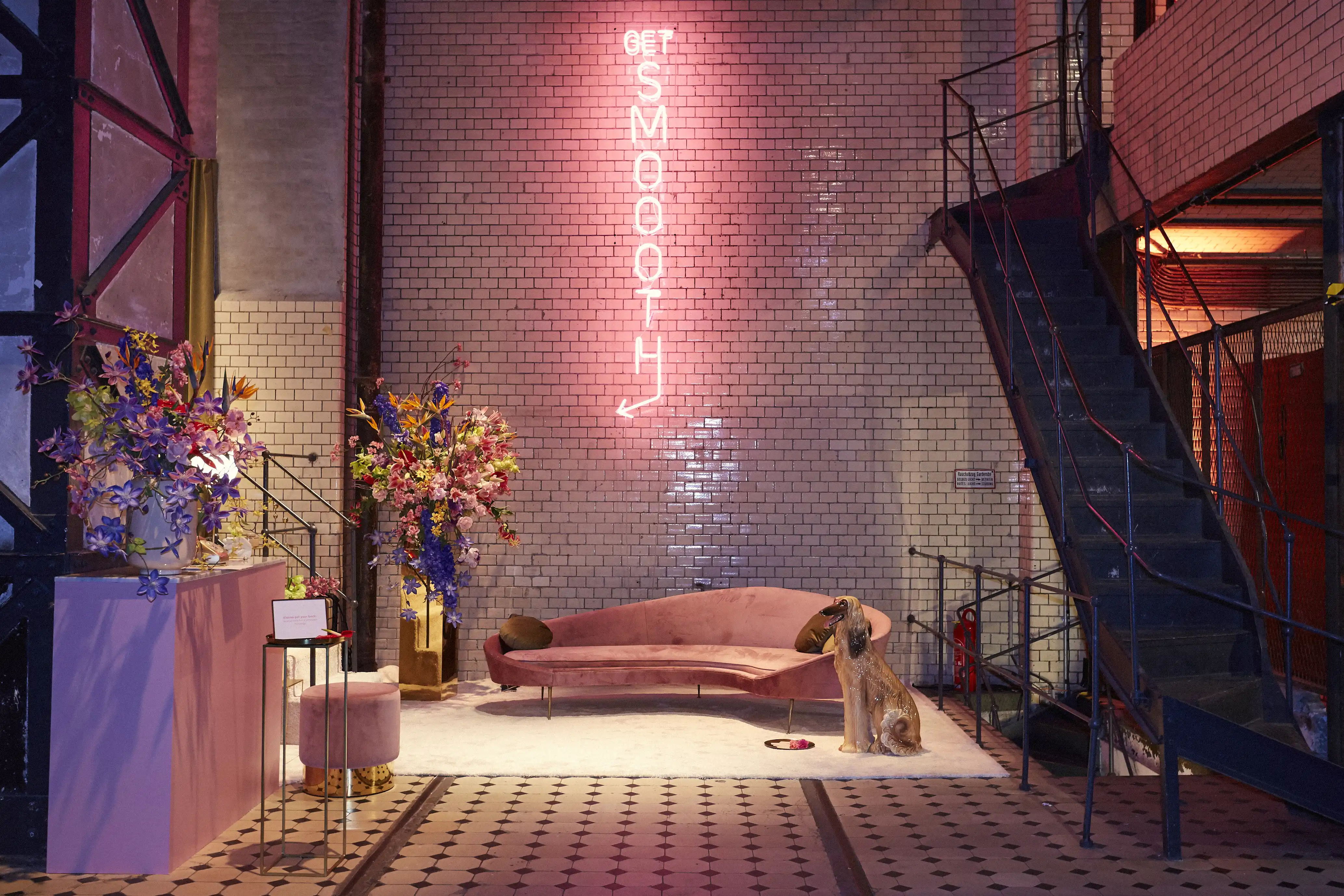 Afghan hound sitting beside a curved pink velvet sofa under the Get Smoooth neon sign on a tiled wall at the Klarna pop-up.