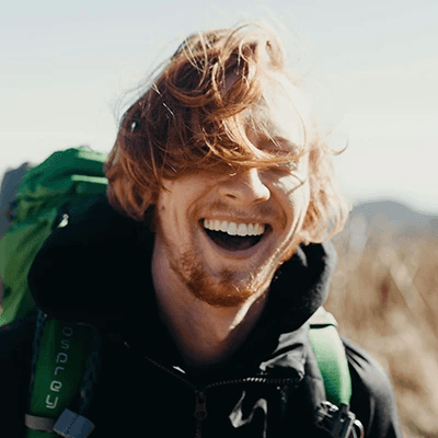 Smiling person with wind-swept hair stands outdoors, wearing a black jacket and green backpack. Bright, sunny day with blurred natural background.